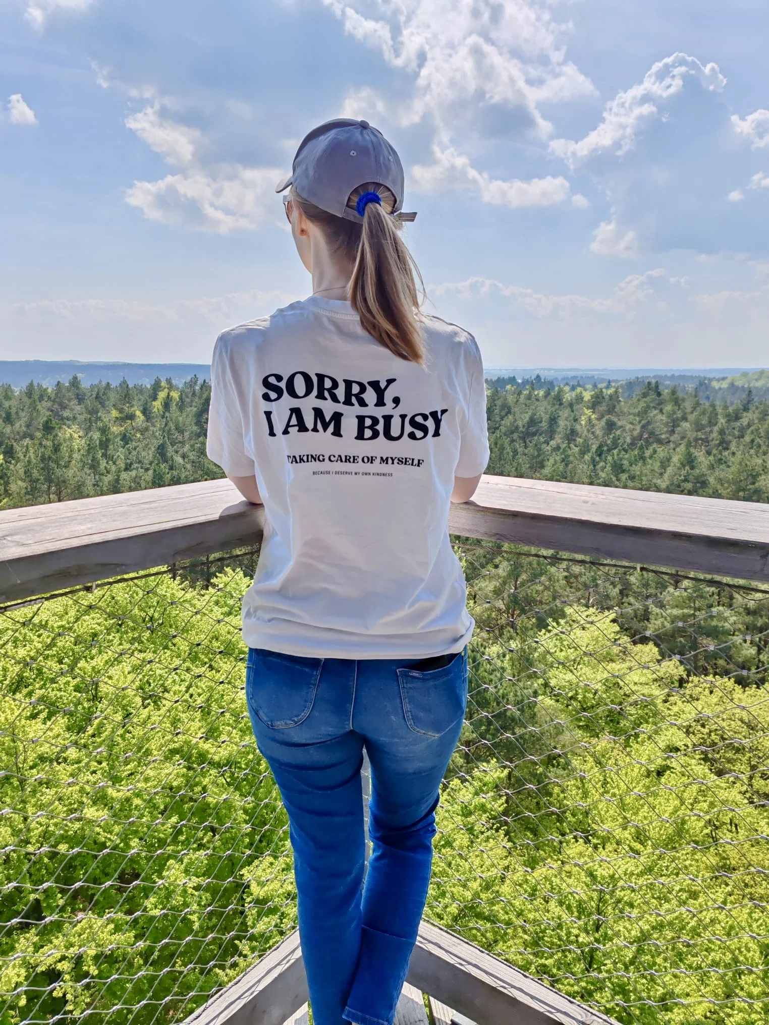 Eine Person mit brauner Haartolle in einem Pony, die einen grauen Baseballkappen trägt, schaut auf eine grüne Waldlandschaft. Sie trägt ein weißes T-Shirt mit schwarzer Schrift, dass lautet: "Sorry, I am busy taking care of myself because I deserve my own kindness". Sie steht auf einer Aussichtsplattform aus Holz, umgeben von einem Zaun aus Draht.