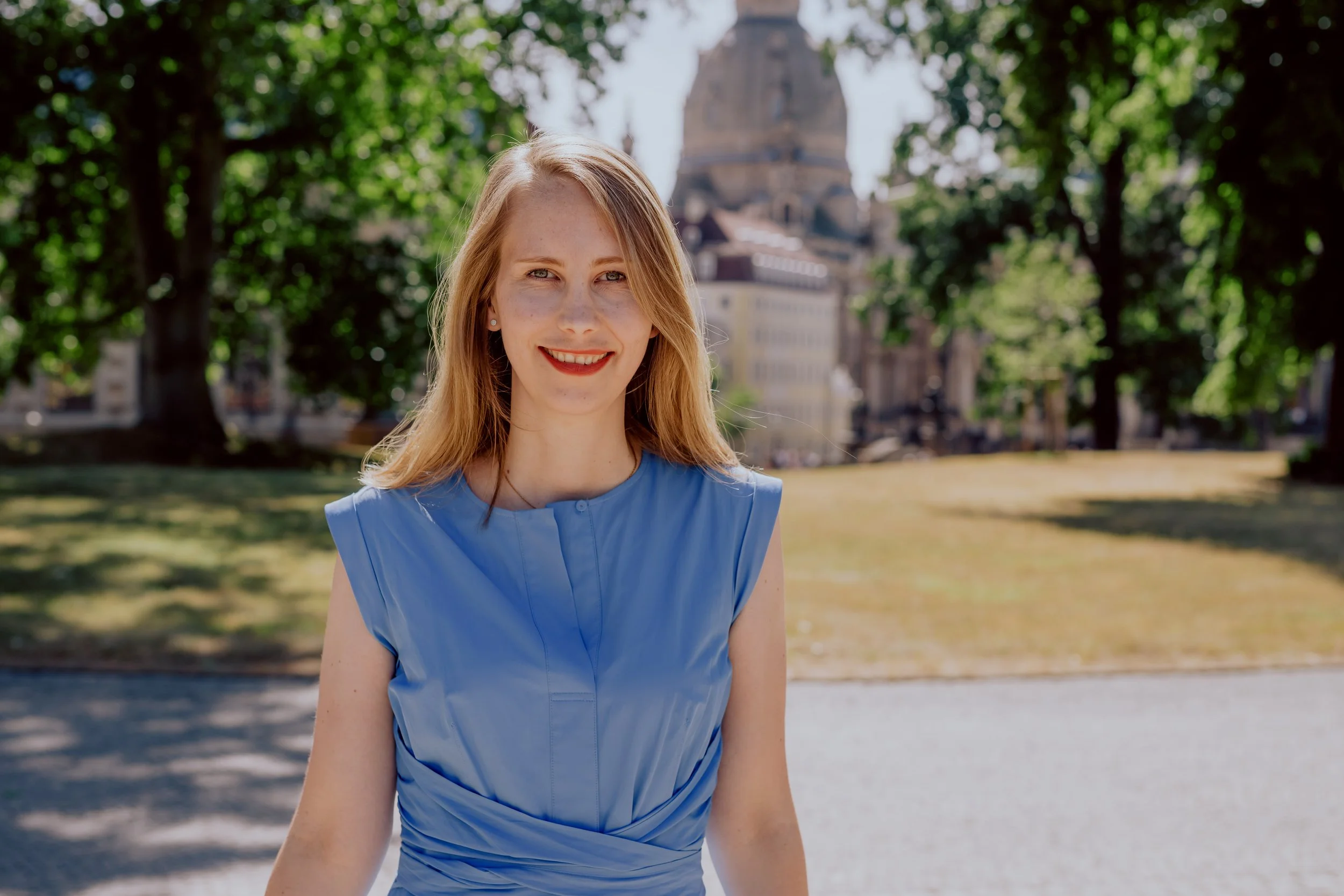 Junge Frau mit blauer Bluse, braune Haare, rote Lippen, vor Kulisse eines Parks mit grünen Bäumen und einer alten Kirche im Hintergrund.