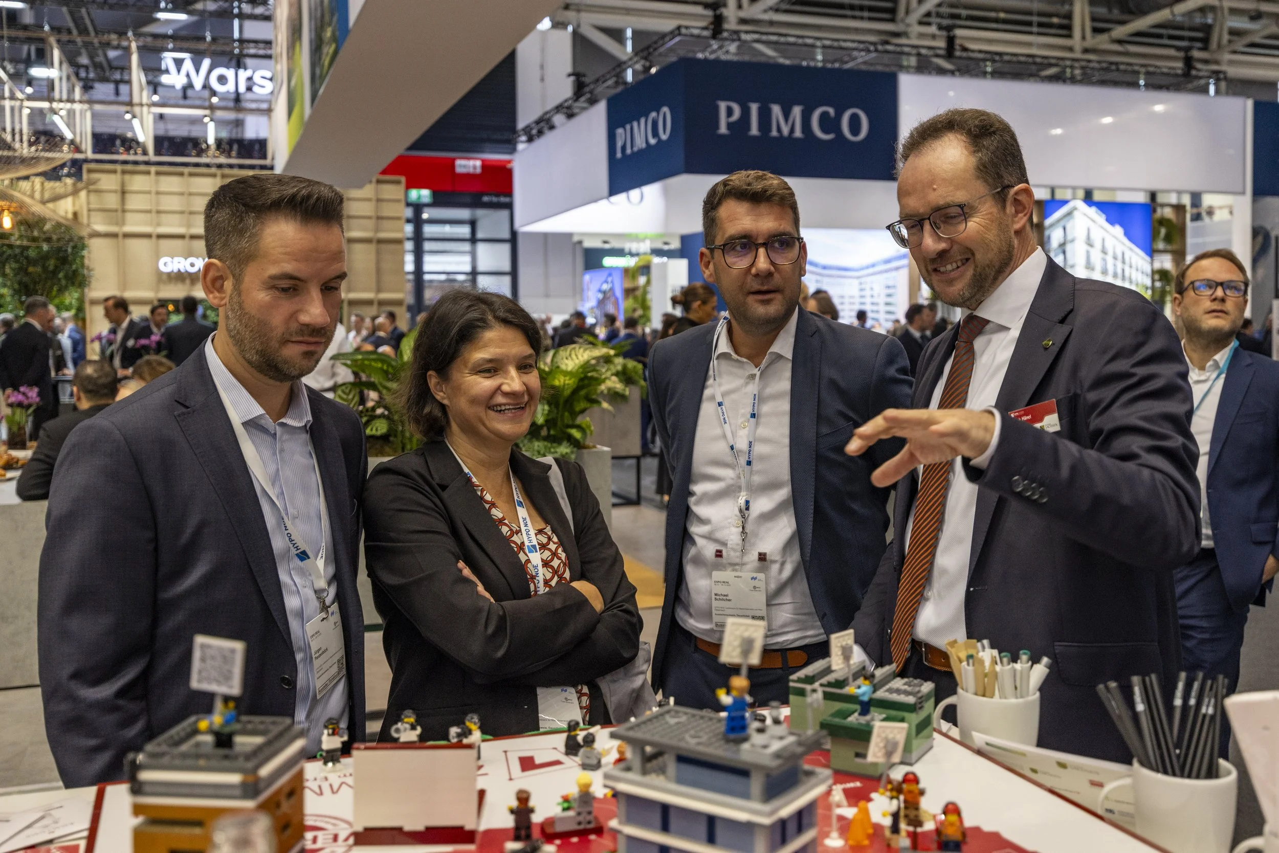 Vier Menschen im Gespräch auf einer Messe, im Hintergrund Stand mit der Beschriftung "PIMCO" und weitere Besucher.