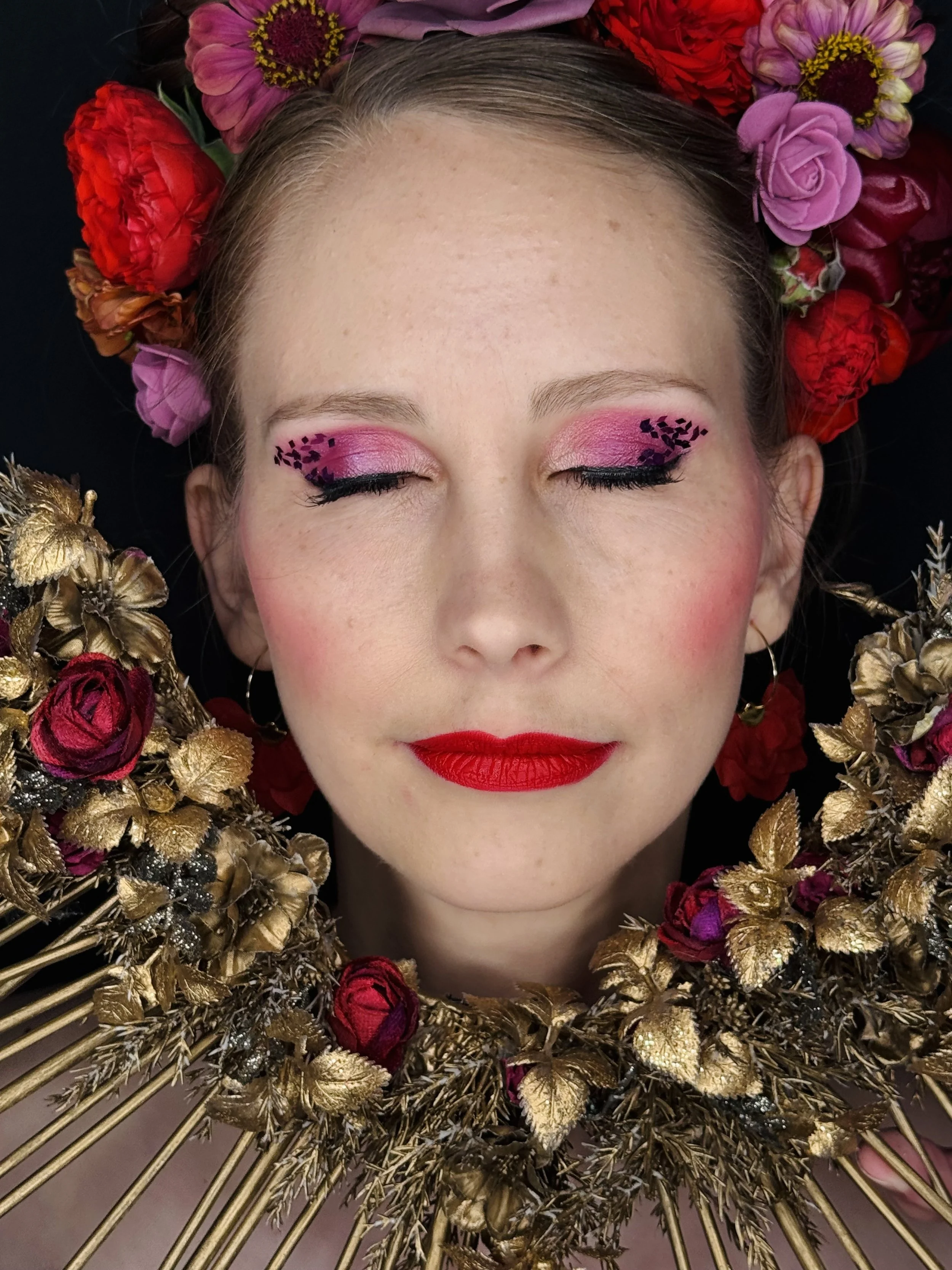 Nahaufnahme eines Gesichts mit pinkem Make-up und roten Lippen, umgeben von Blumen: Rosen, Hortensien und weiteren blühenden Pflanzen in einem goldfarbenen Rahmen.