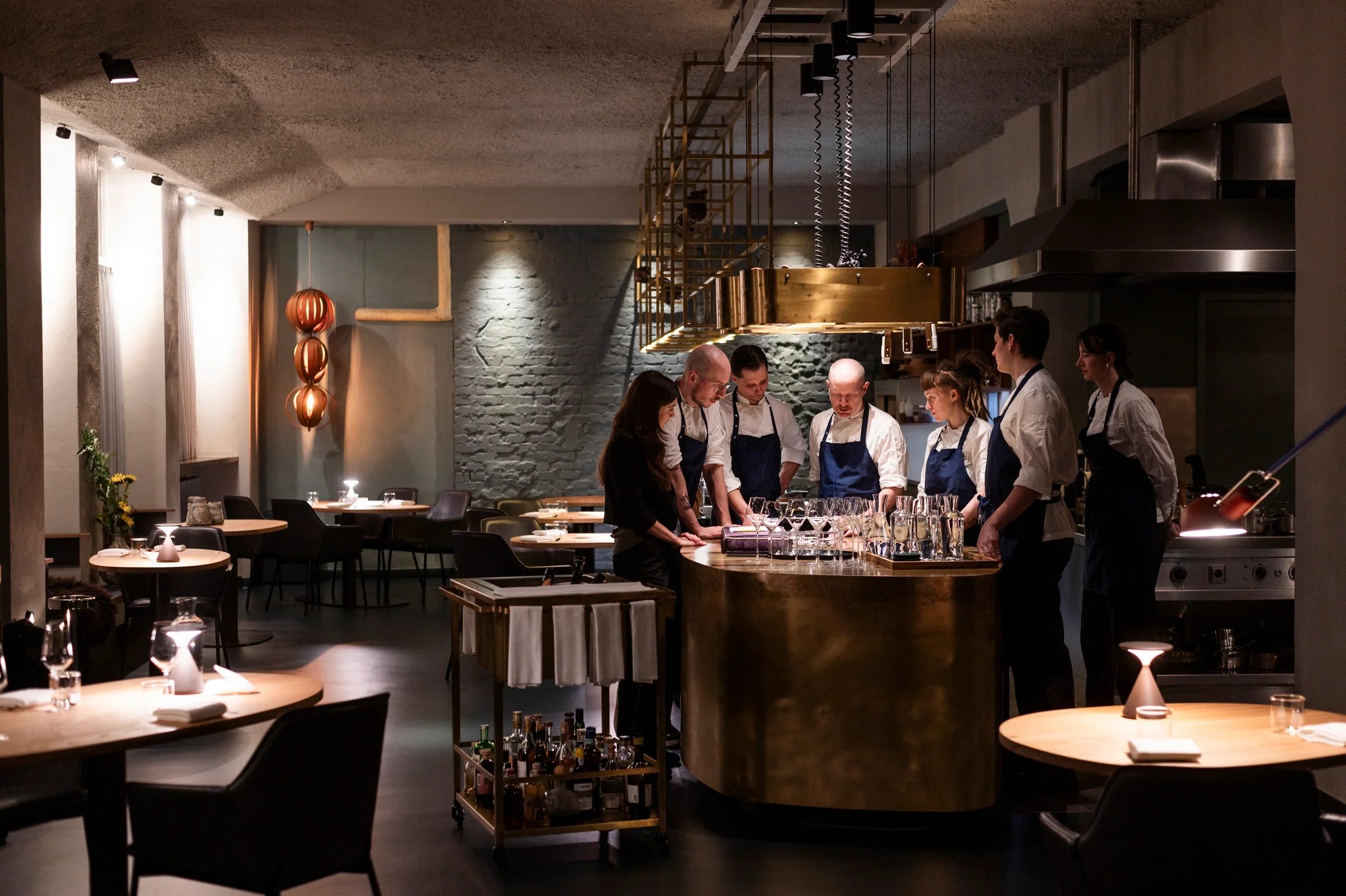Küche in einem Restaurant mit mehreren Köchen, die an einem runden Thekenbereich arbeiten, umgeben von dunkler Inneneinrichtung, Beleuchtung und Dekoration im Hintergrund.