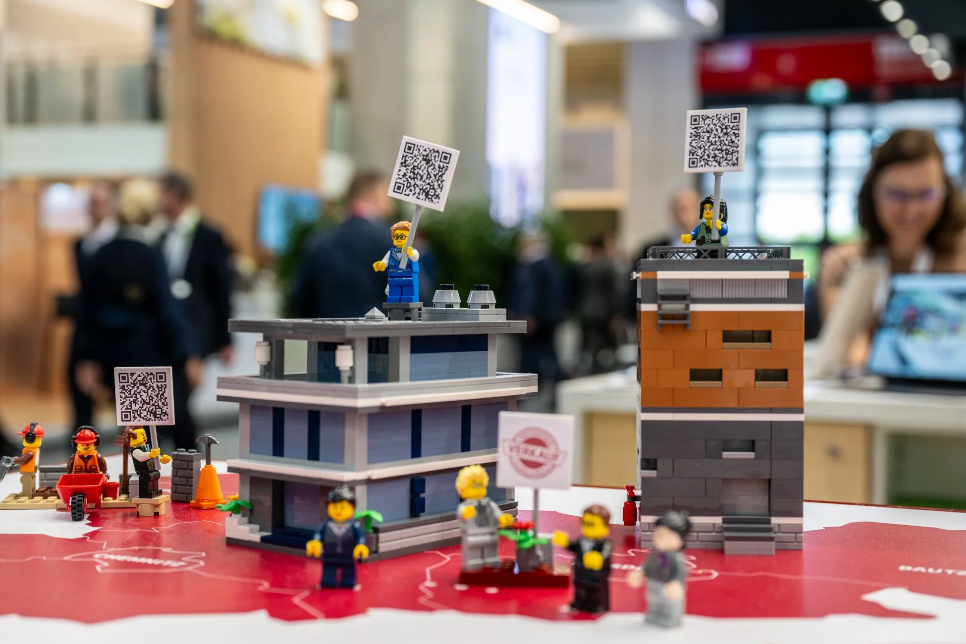 Diorama aus LEGO-Steinen mit mehreren Szenen und Figuren, QR-Codes auf Ständern, im Hintergrund Menschen in einem Ausstellungsraum.