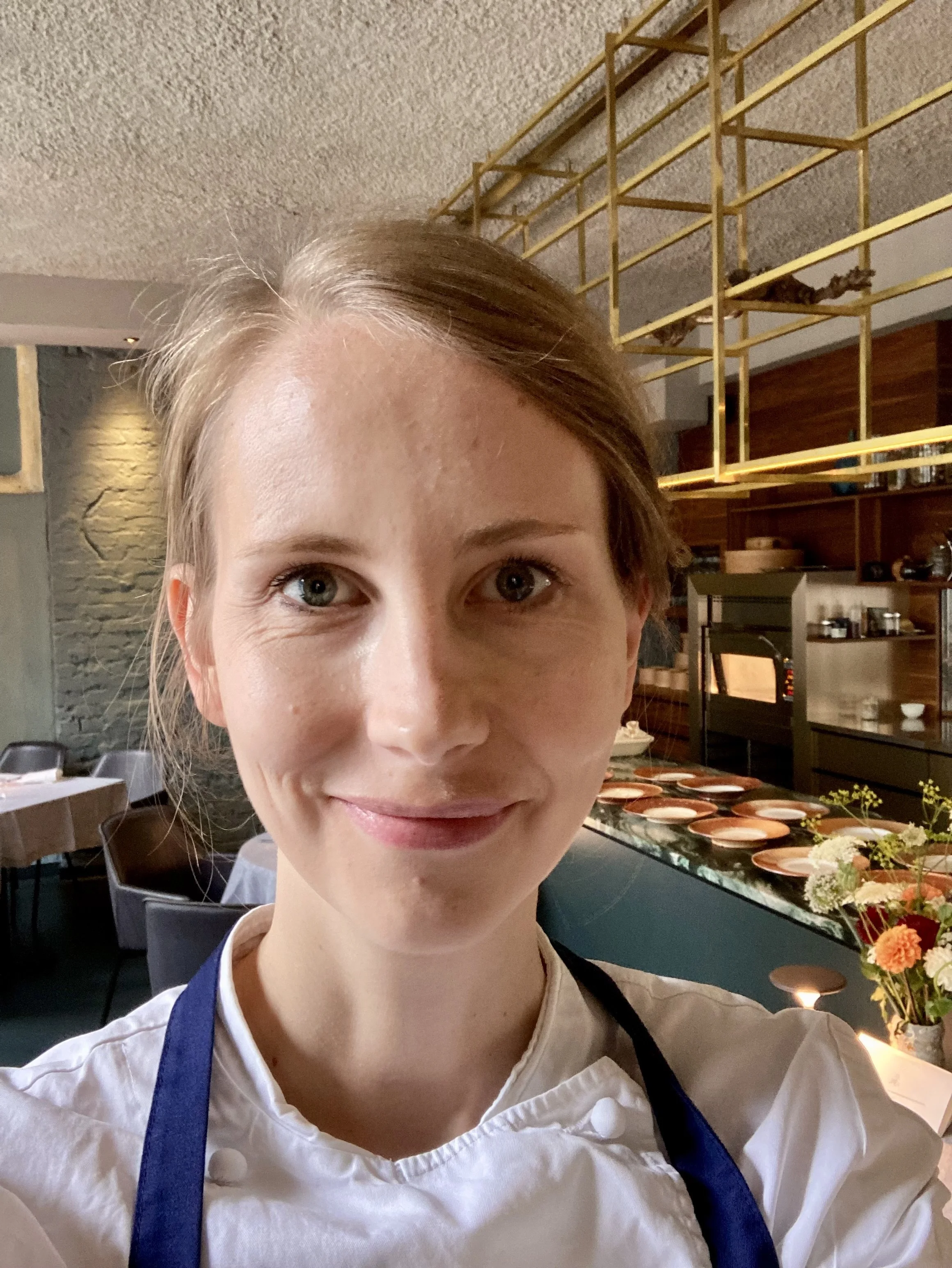Ein Lächeln einer jungen Frau, die in einem Restaurant oder Küchenumfeld steht, trägt eine weiße Kochjacke und eine blaue Schürze. Im Hintergrund sind Tische, Geschirr und ein Buffet mit Tellern und Blumen zu sehen.