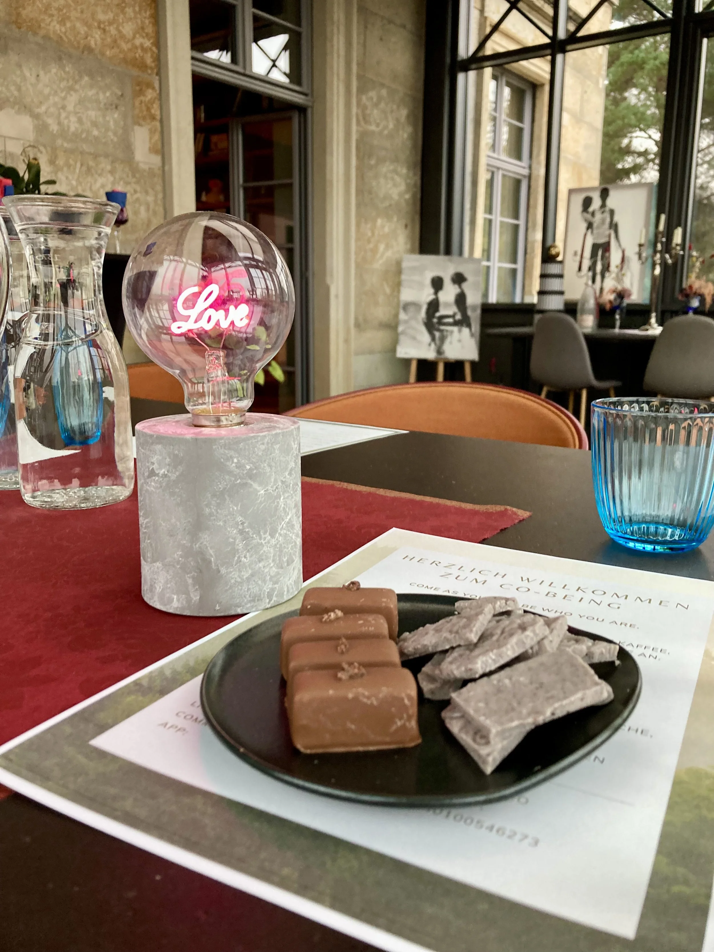 Ein Teller mit Schokoladentafeln und getrockneten Blättern auf einem Tisch in einem Café, im Hintergrund sind Fenster und Wanddekorationen sichtbar.
