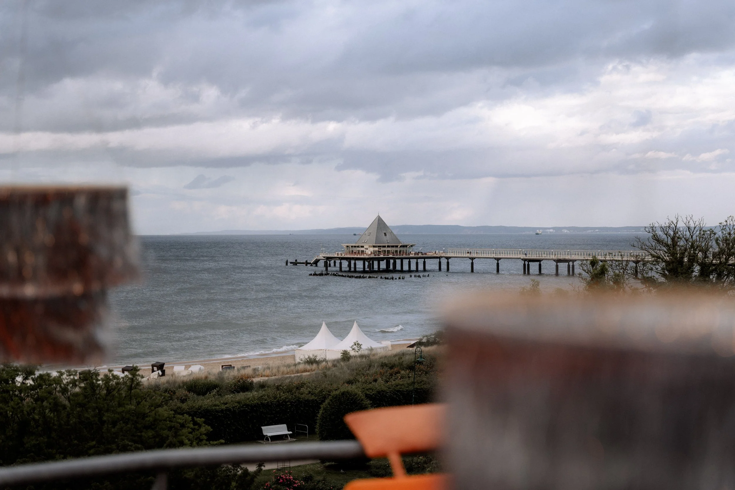 Blick auf den Strand mit einem Pier und einer Gebäudestruktur im Wasser, umgeben von dunklen Wolken am Himmel, eingerahmt von unscharfen Elementen im Vordergrund.