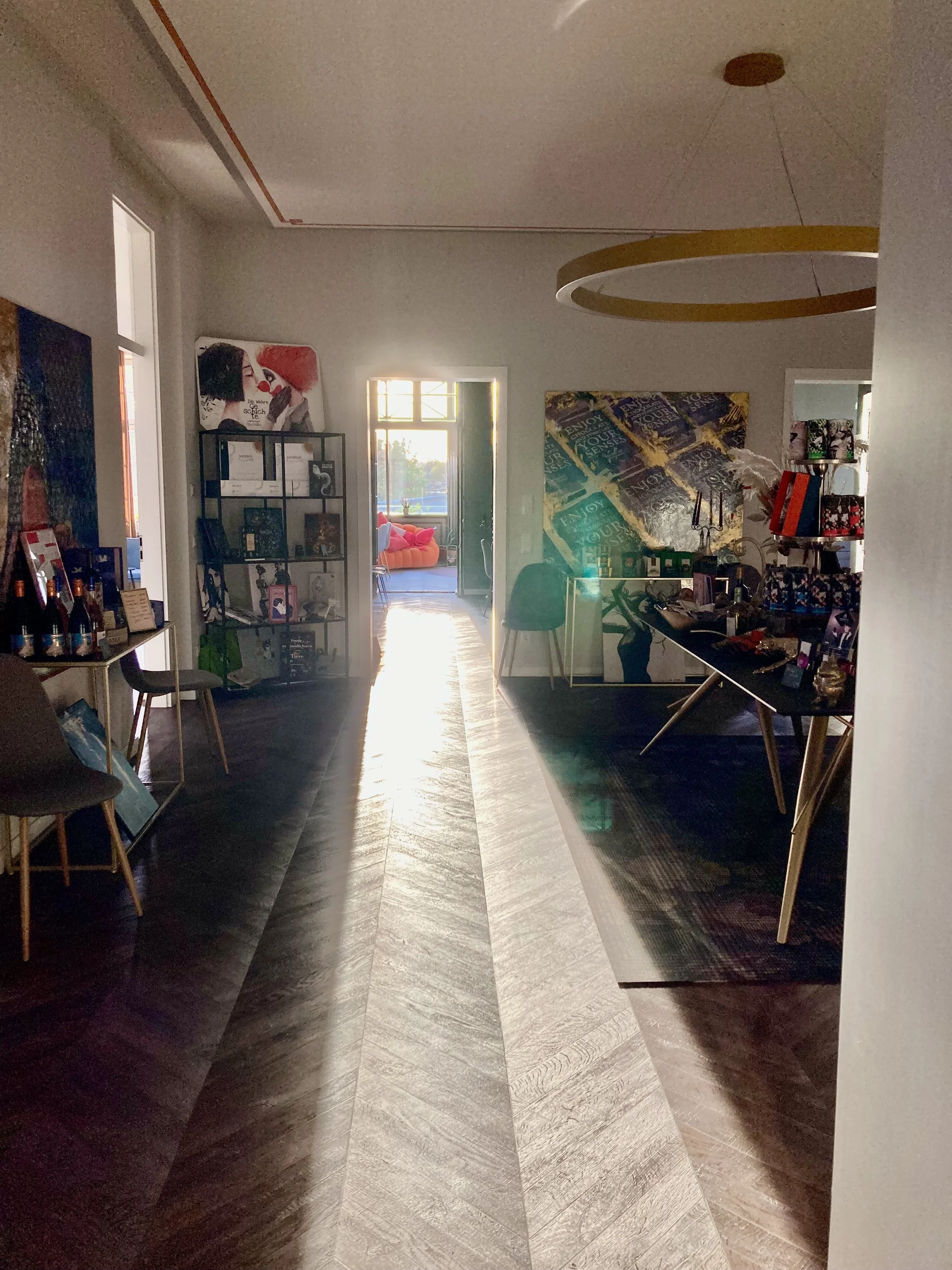 Innenaufnahme eines Wohnzimmers mit dunklem Dielenboden, weißem Dach und mehreren Design-Wandbildern. Es gibt einen offenen Durchgang zum Balkon mit Blick nach draußen, viele Möbel und Dekorationsgegenstände.