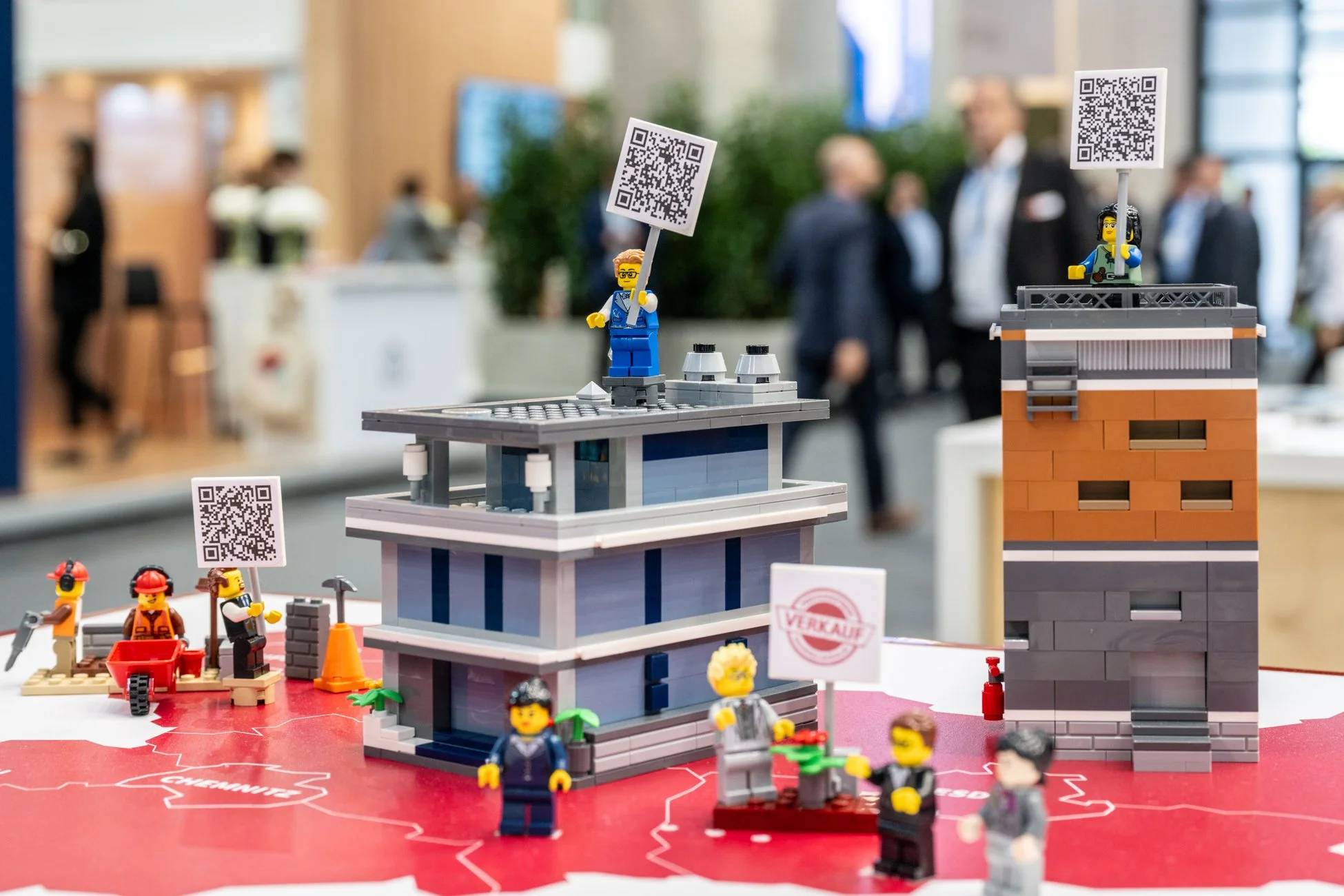 Lego-Szene mit Bauarbeitern, Gebäuden und QR-Codes auf einer Messe oder Ausstellung.
