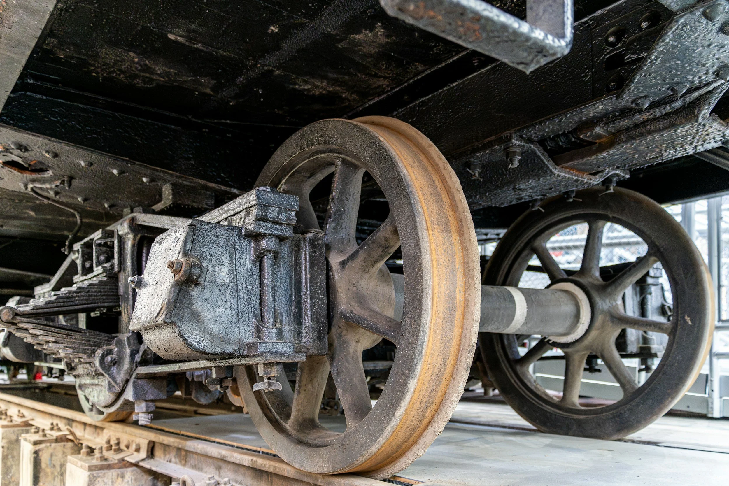 Underneath view of a train wheel and track, showing the rusted wheel and mechanical parts with oil and dirt, on a wooden pallet.