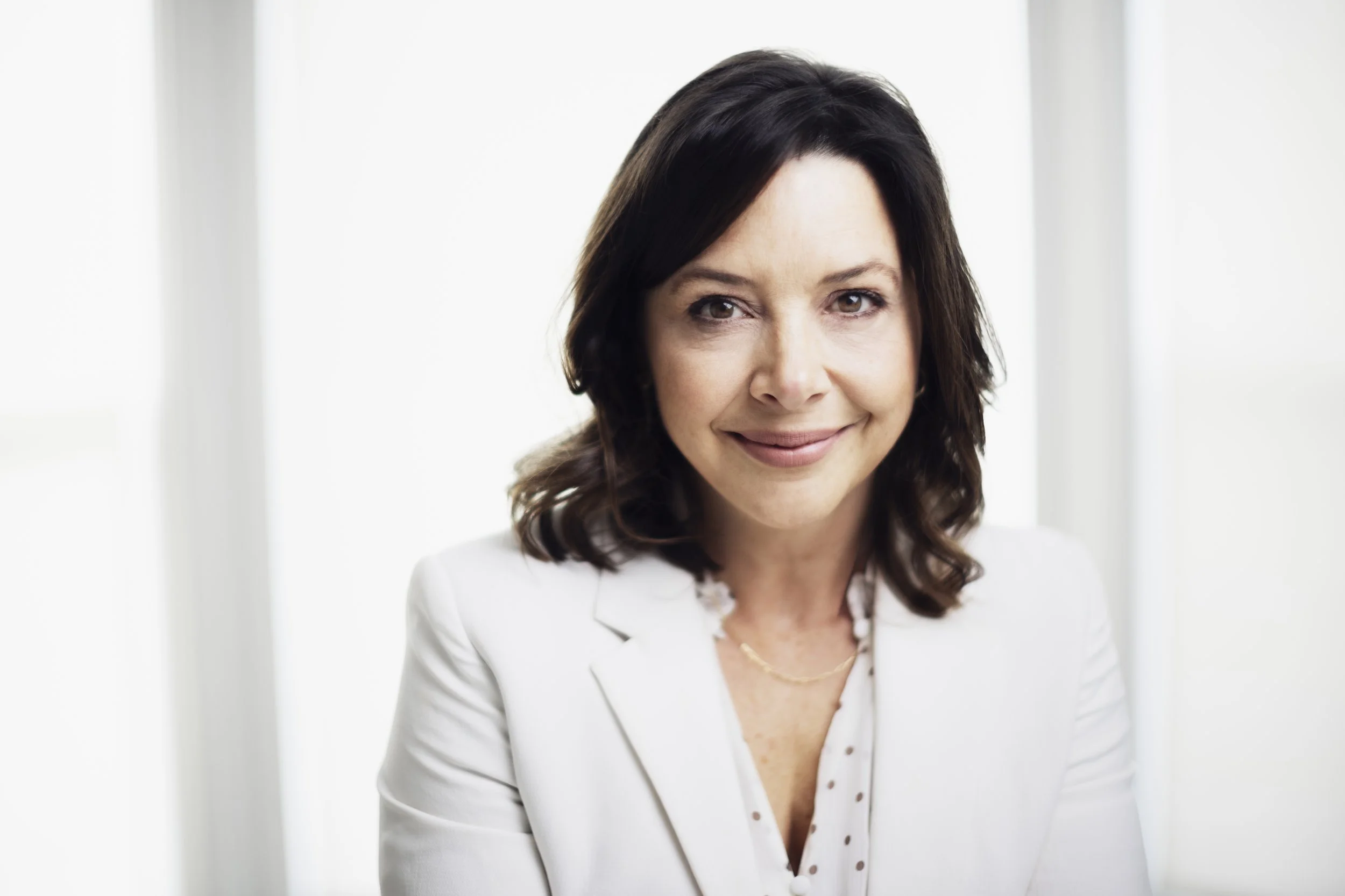 Professional woman with dark brown, shoulder-length hair, wearing a white blazer and polka dot blouse, smiling at the camera indoors with bright natural light.