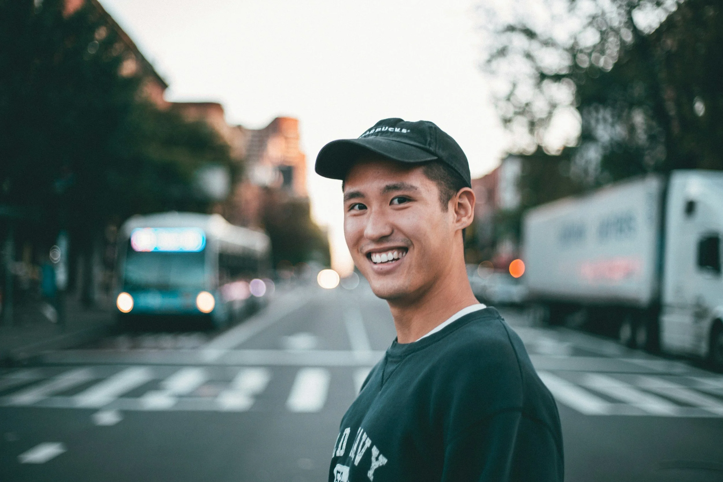 A young man smiling and looking at the camera standing on a city street with a crosswalk in the foreground, blurred bus, trucks, and buildings in the background during dusk or evening.