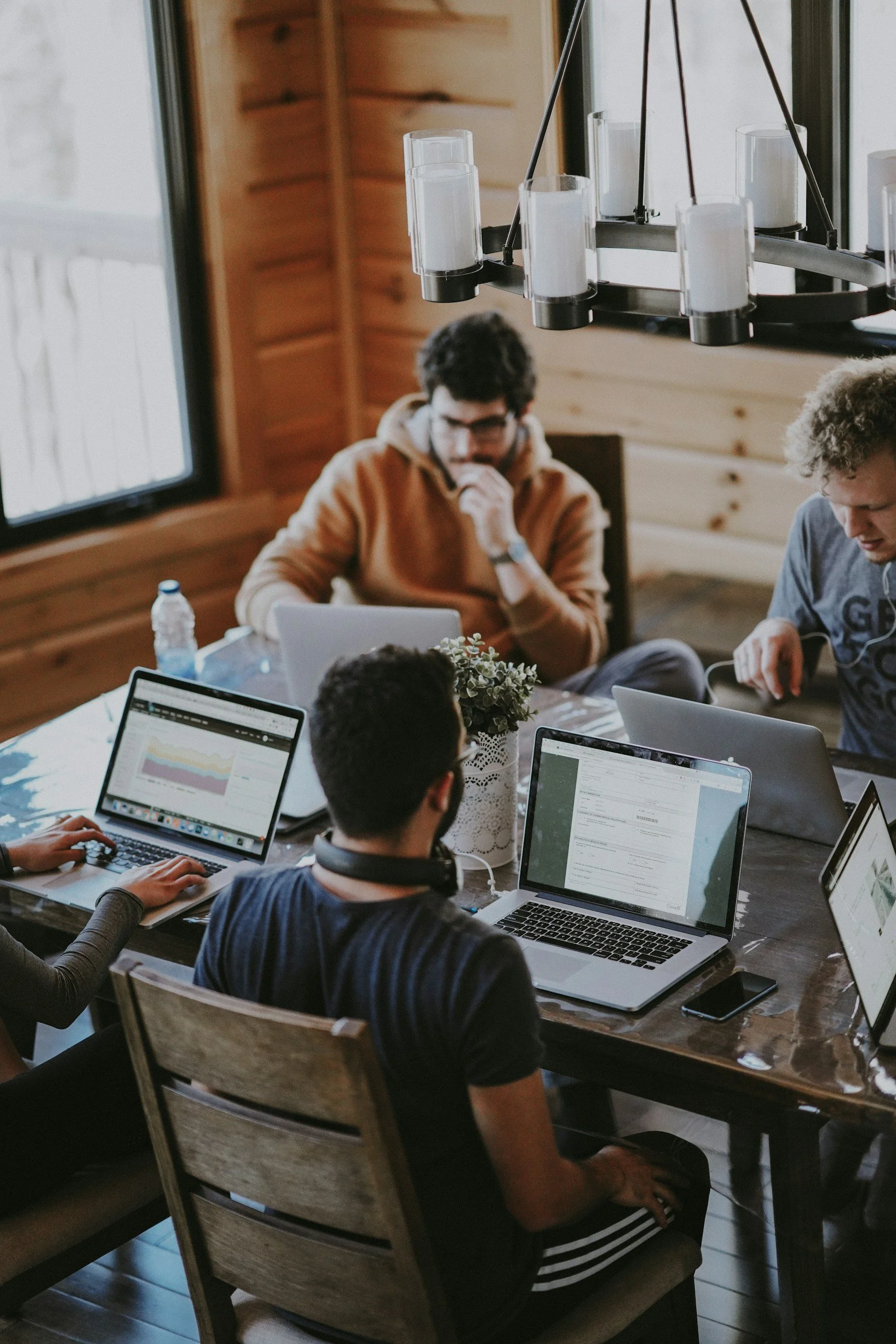 A group of four young men working on laptops at a wooden table in a cozy, rustic room with natural light.