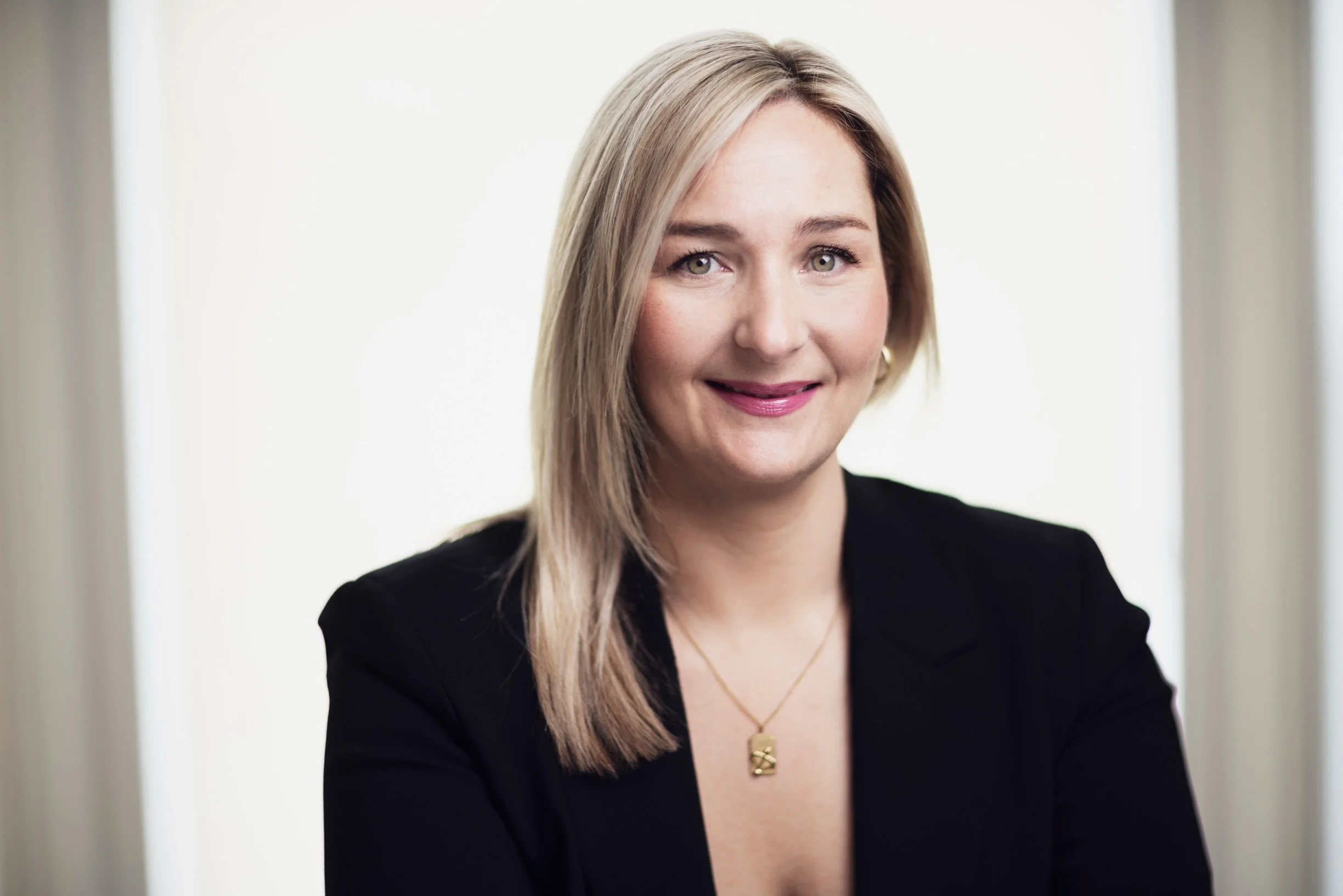 A woman with blonde hair wearing a black blazer and gold jewelry, smiling and looking at the camera.