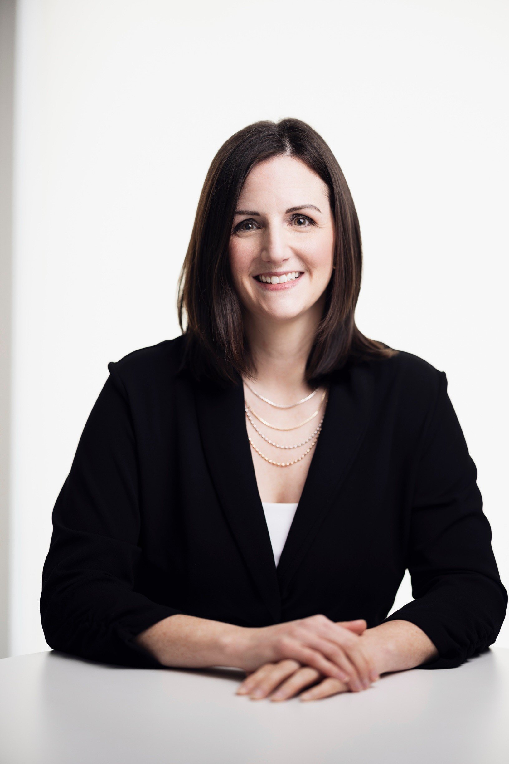 Portrait of a woman with shoulder-length dark brown hair, wearing a black blazer and layered jewelry, sitting at a white table against a plain white background.