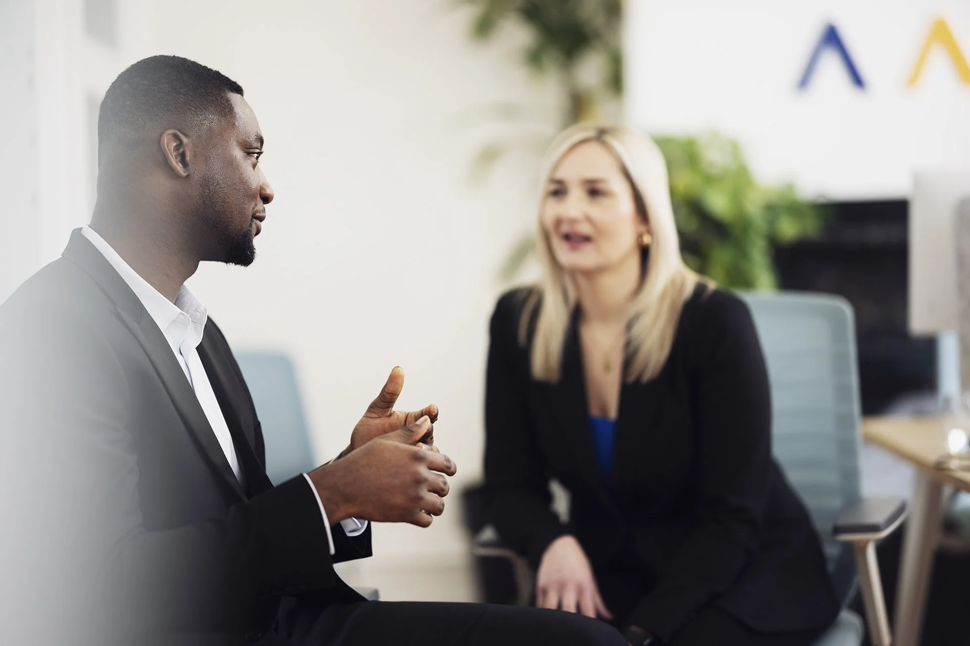 A man and woman in a professional office setting engaged in conversation. The man, wearing a black suit and white shirt, appears to be explaining something with hand gestures. The woman, dressed in black, listens attentively. Background includes office furniture and plants.
