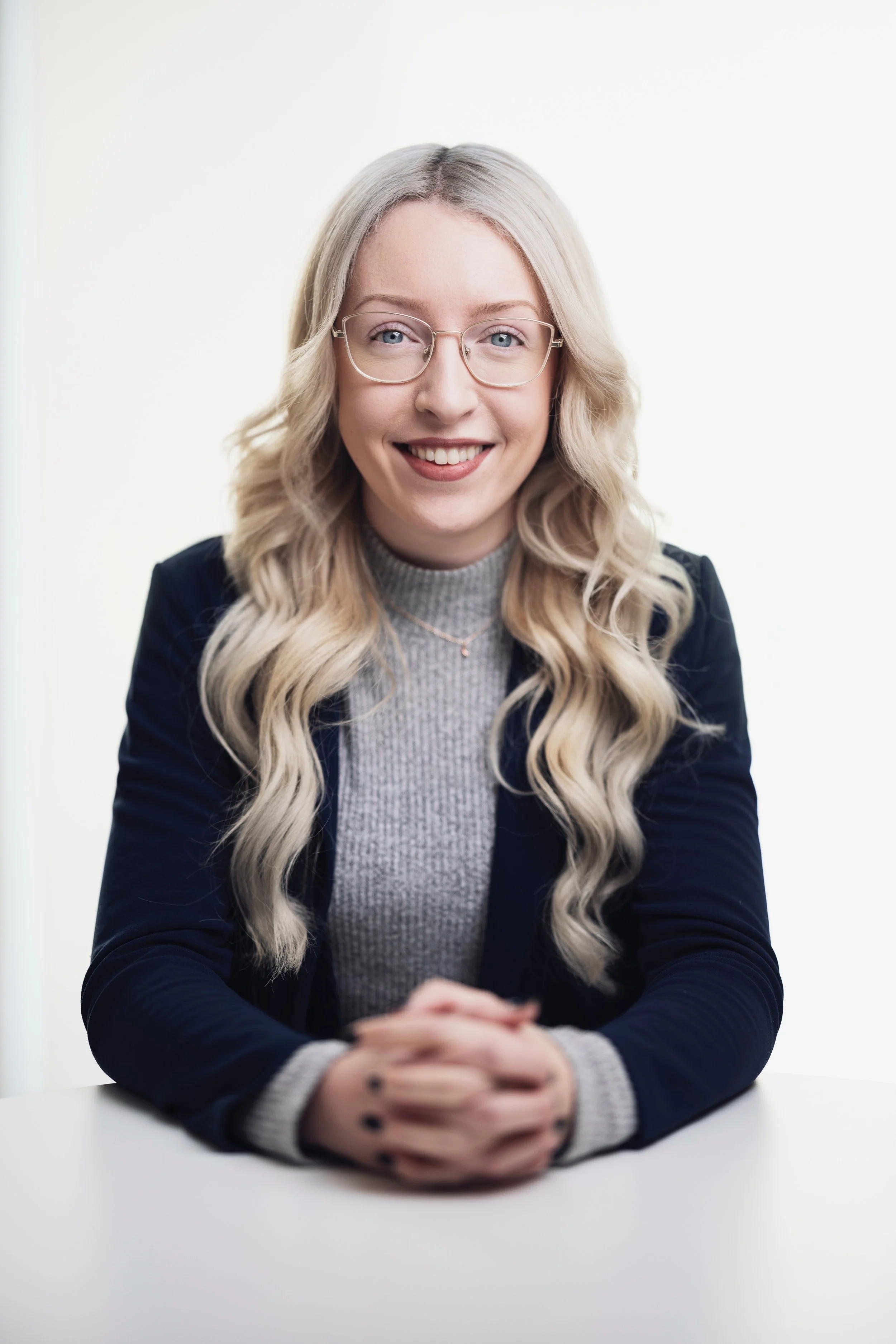 A young woman with long, wavy blonde hair, wearing glasses, a gray turtleneck, and a navy blazer, sitting at a table with hands clasped, smiling at the camera.