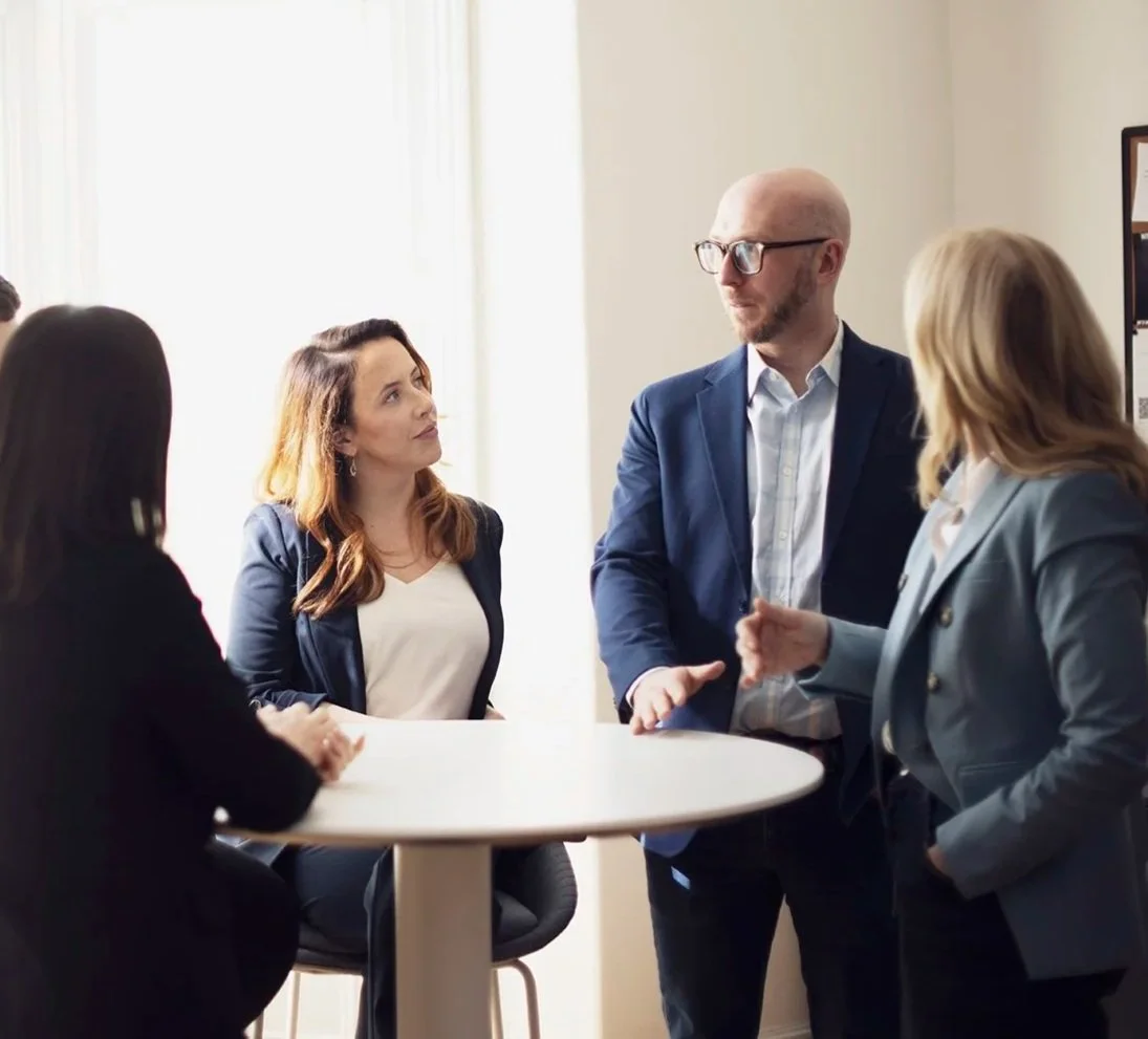 Four business professionals having a discussion in an office, three women and one man, with the man standing and the women sitting around a round table.