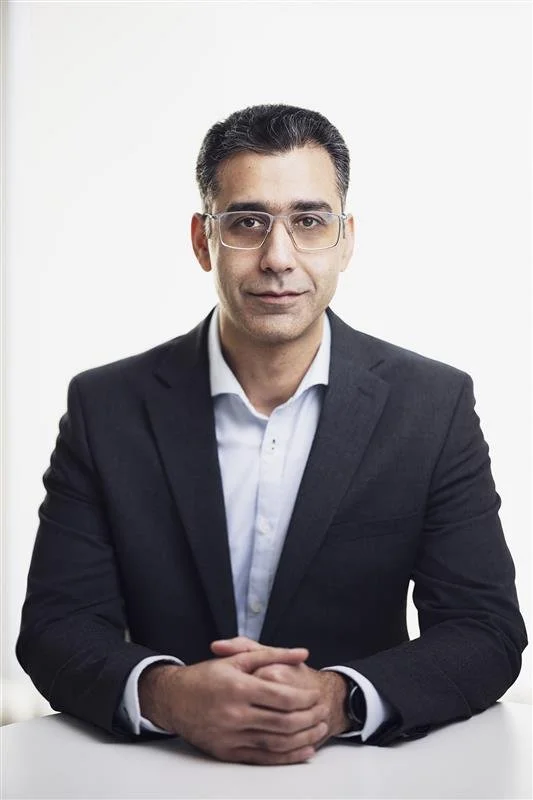 A professional man with glasses dressed in a dark suit and white shirt, sitting at a white table against a plain white background.