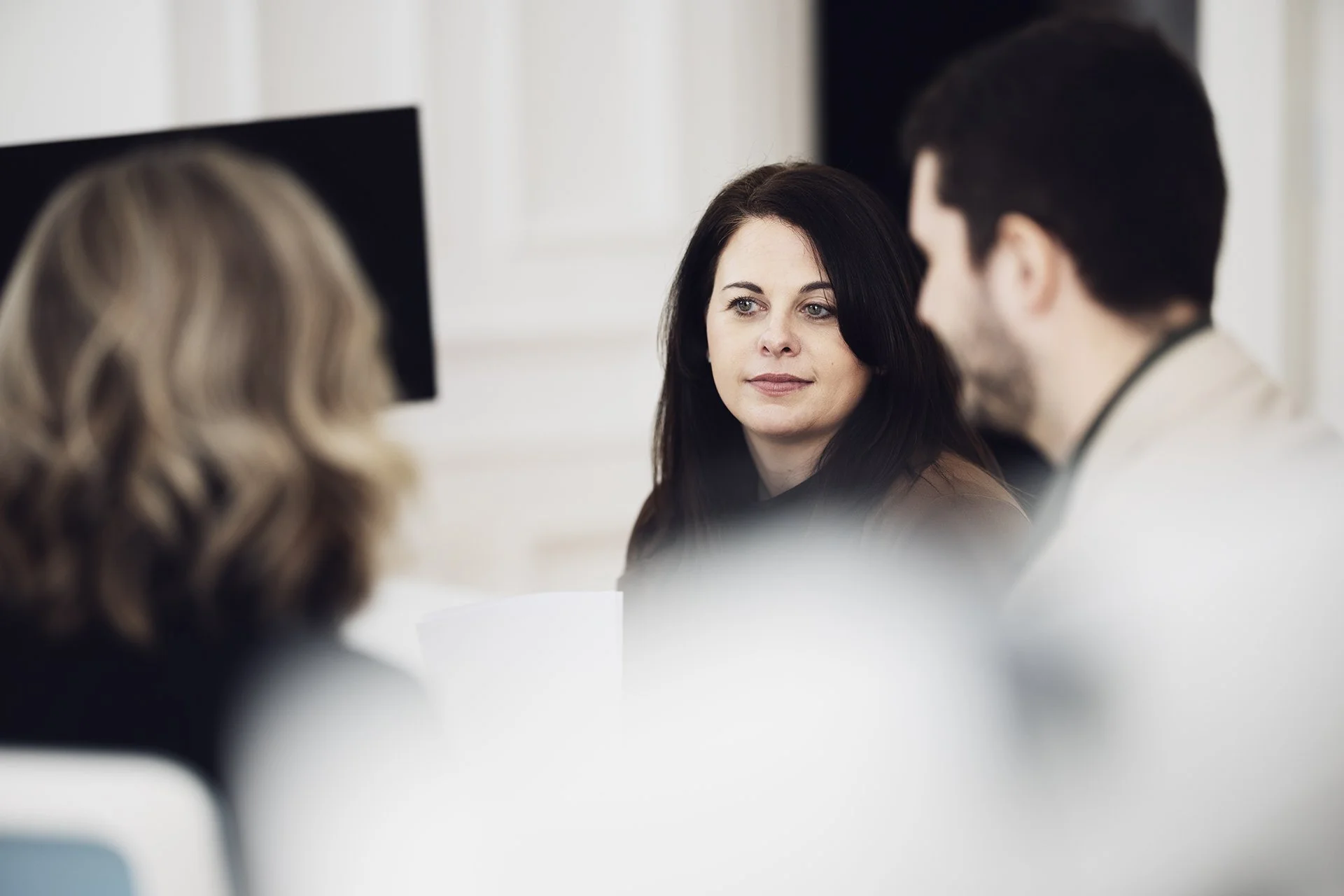 A woman with dark hair looking attentively at a man with dark hair and beard, both engaged in conversation with another woman whose blonde hair is visible, in a bright room.