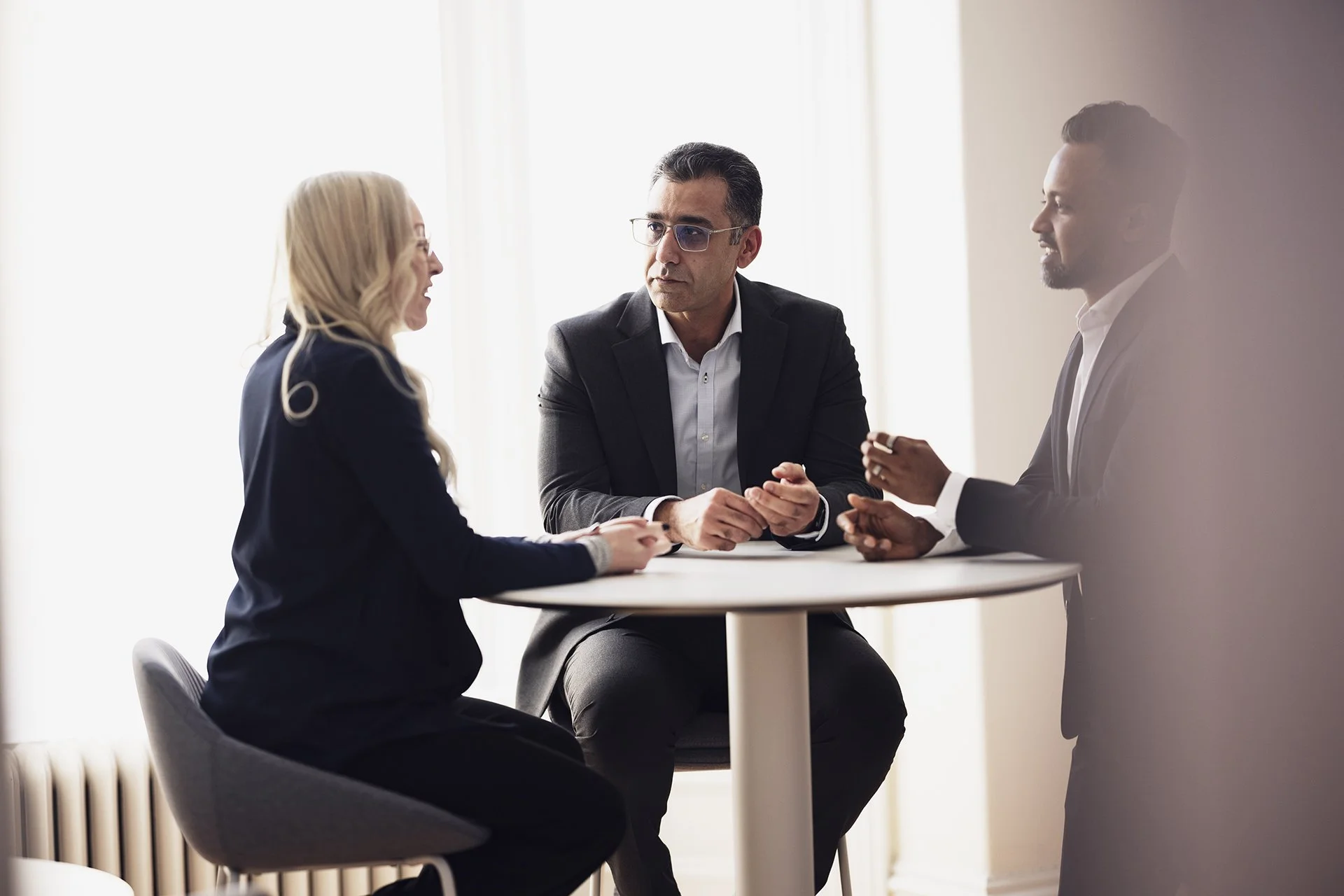 Three professionals, two men and one woman, engaged in a discussion around a small round table in an office with bright natural light.