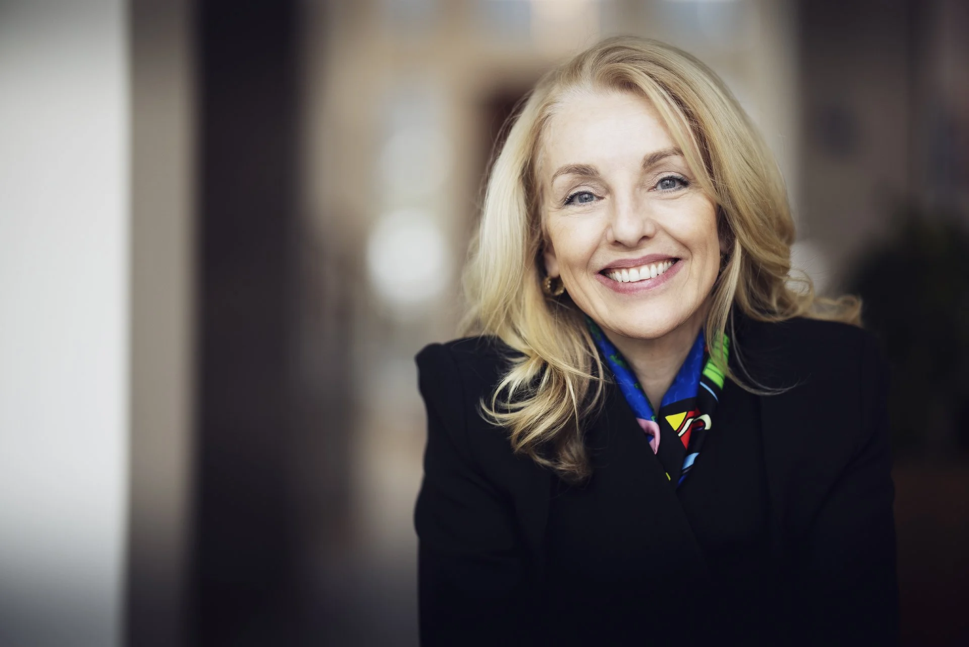 A smiling blonde woman wearing a black blazer and colorful scarf, with a blurred background.