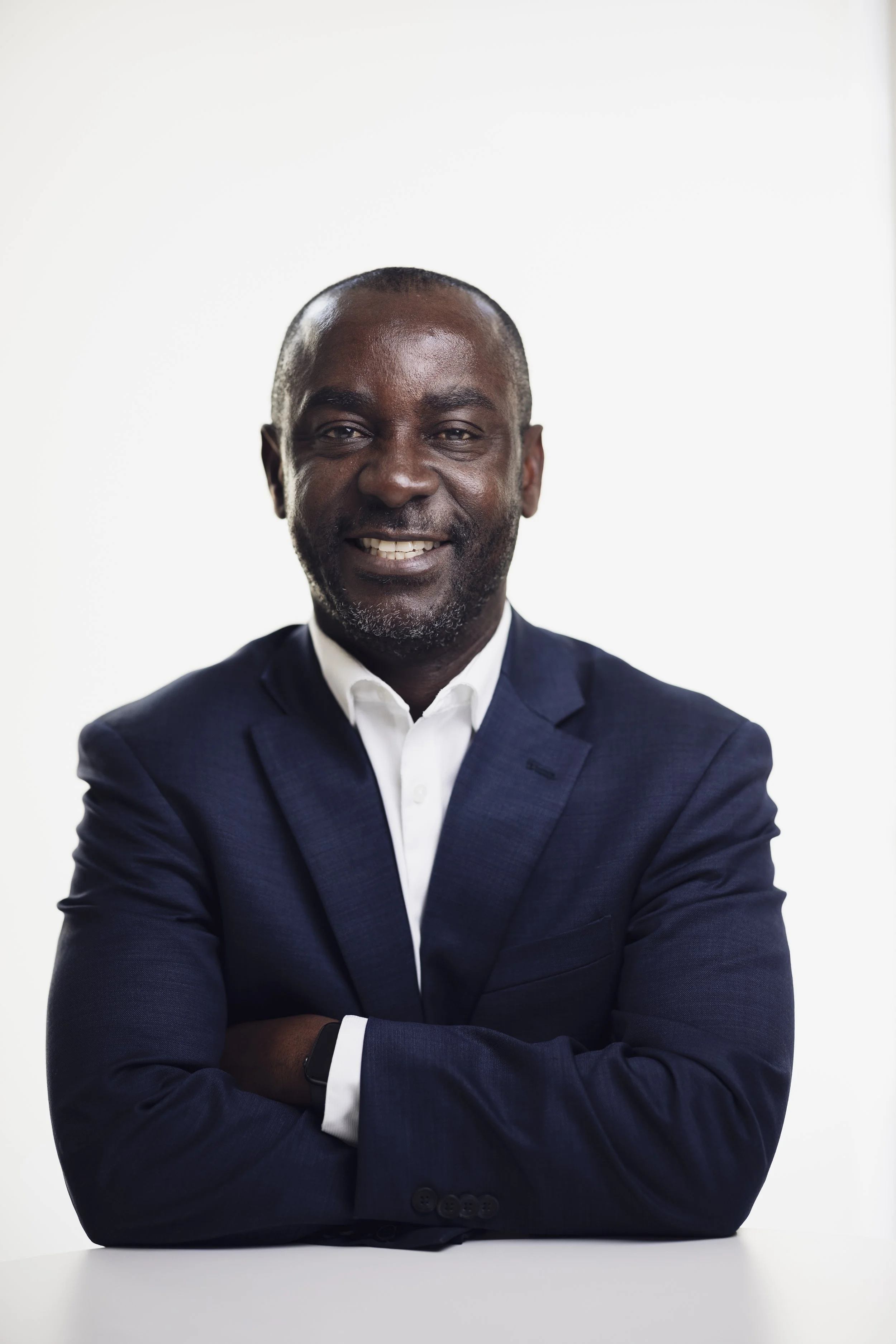 Portrait of a middle-aged African American man in a navy suit and white shirt smiling with arms crossed against a plain white background.