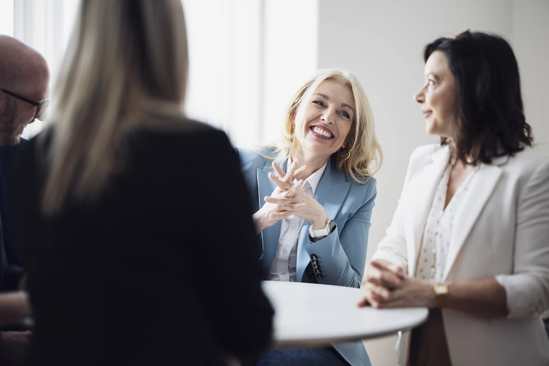Four adults sitting around a white table, engaged in conversation, with a woman with blonde hair smiling and talking to a woman with dark hair.