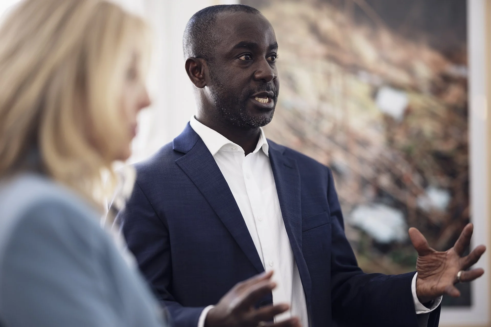 A man in a navy suit and white shirt speaking, with a blurred woman in the foreground listening, in front of an abstract painting.