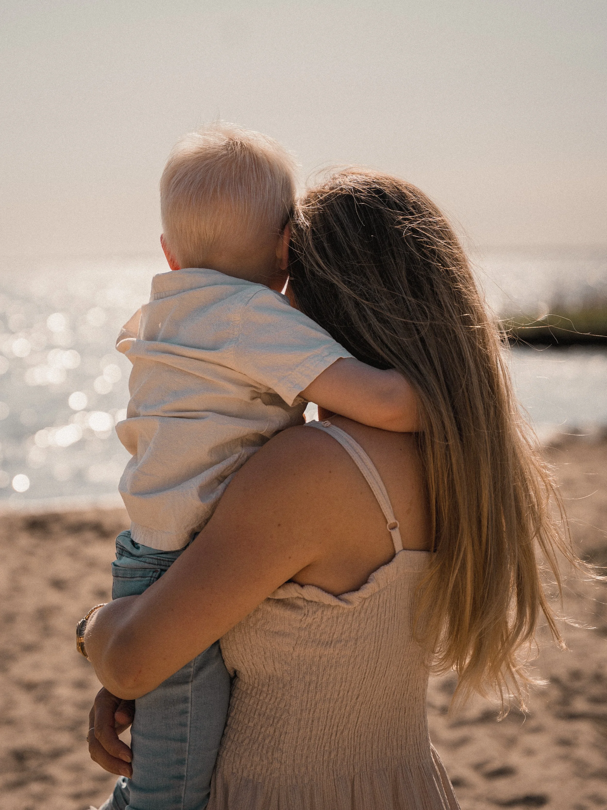 Een vrouw houdt een jonge kind in haar armen op een strand, terwijl ze naar de oceaan kijkt.