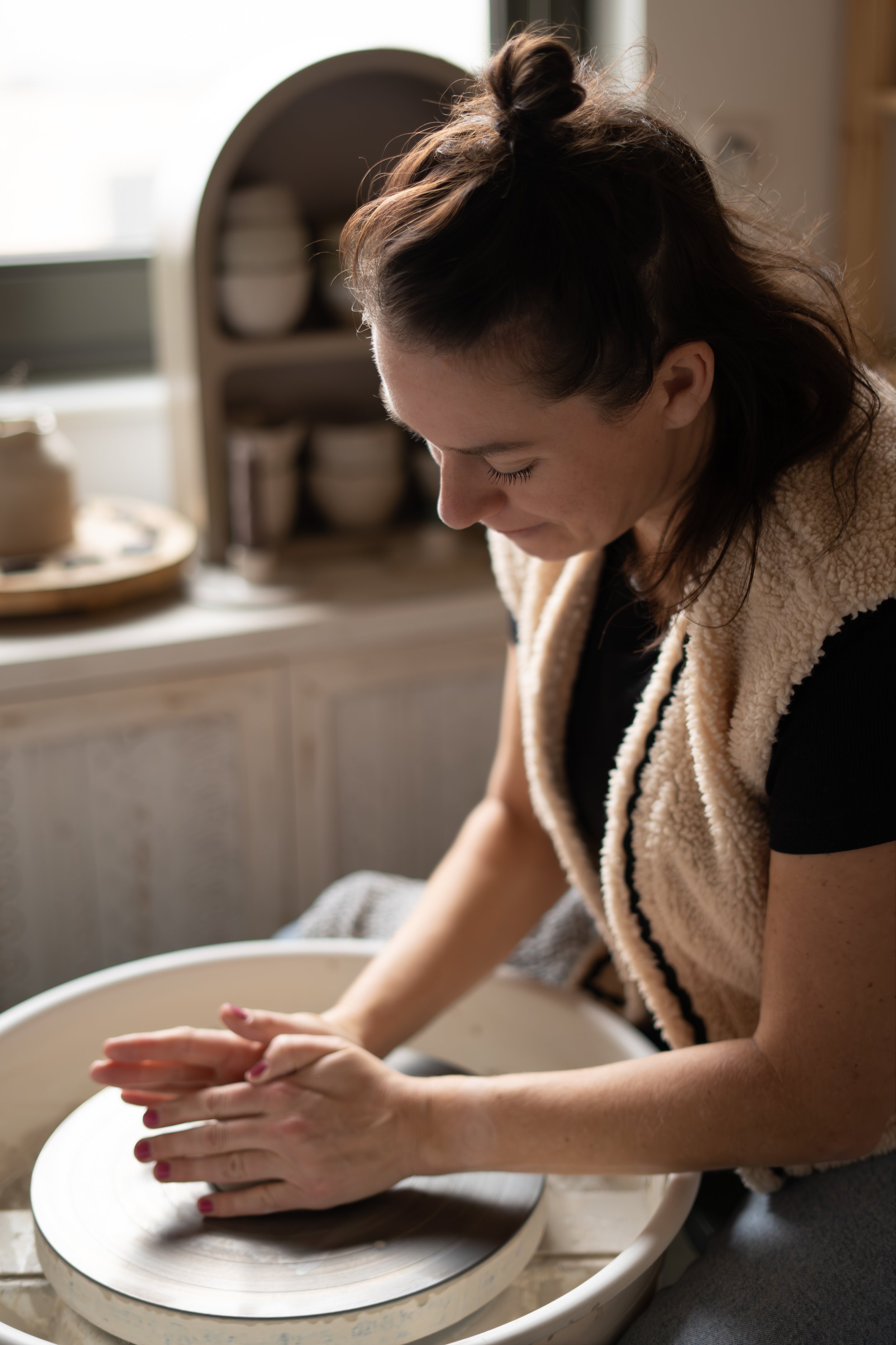 Vrouw zit op een beige gedraaide stoel met haar benen gekruist en gebruikt een laptop, in een lichte kamer met grote witte gordijnen en een groen plant.