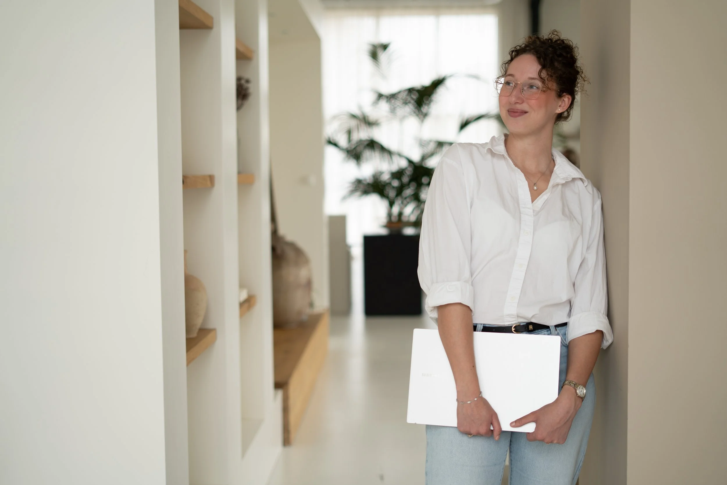 Vrouw met kort krullend haar, bril en een witte blouse die in een huis staat, terwijl ze een laptop vasthoudt.