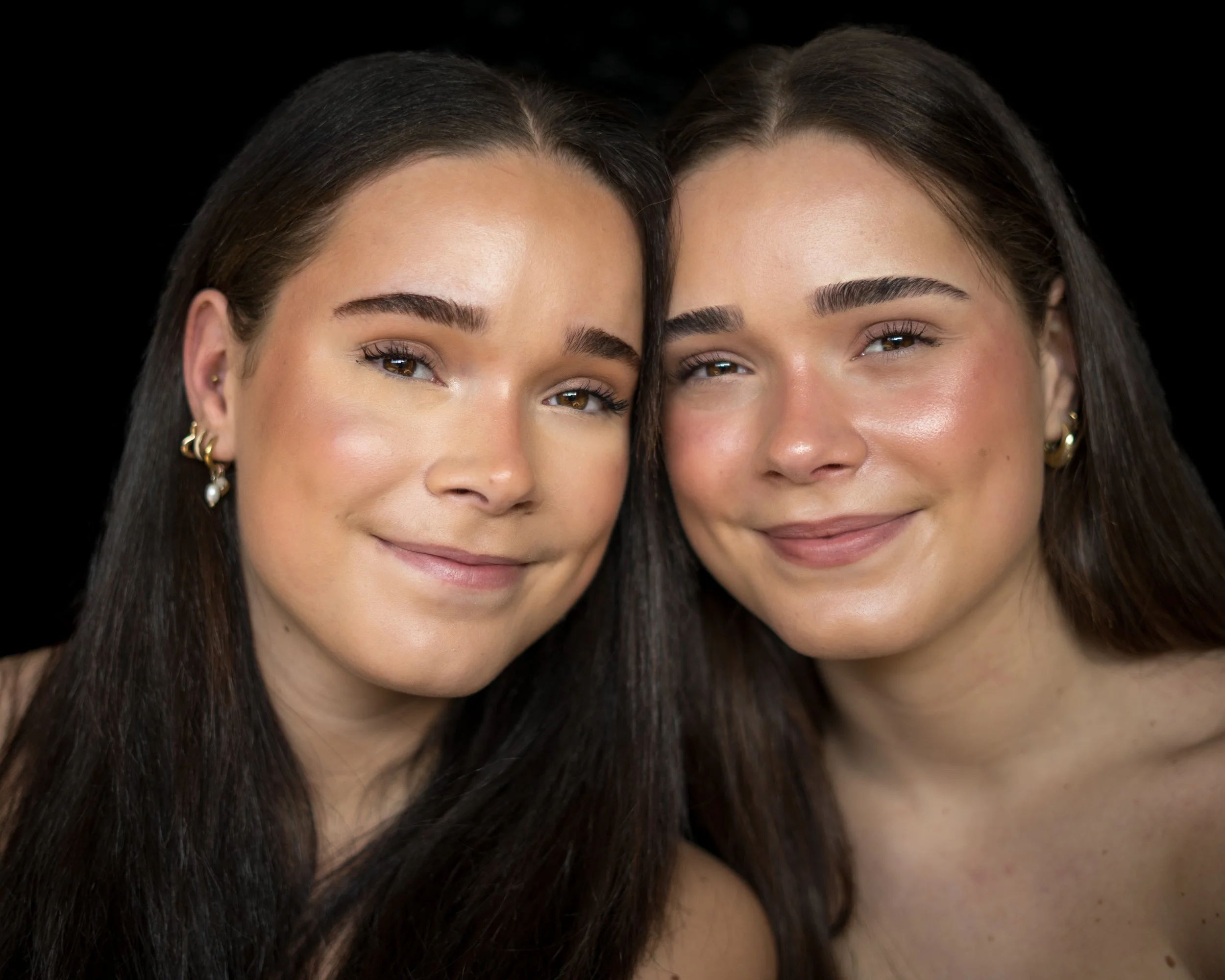 Twee jonge vrouwen met donkere haar, die dicht bij elkaar poseren op een zwarte achtergrond, glimlachend en met casual make-up en goudkleurige oorbellen.