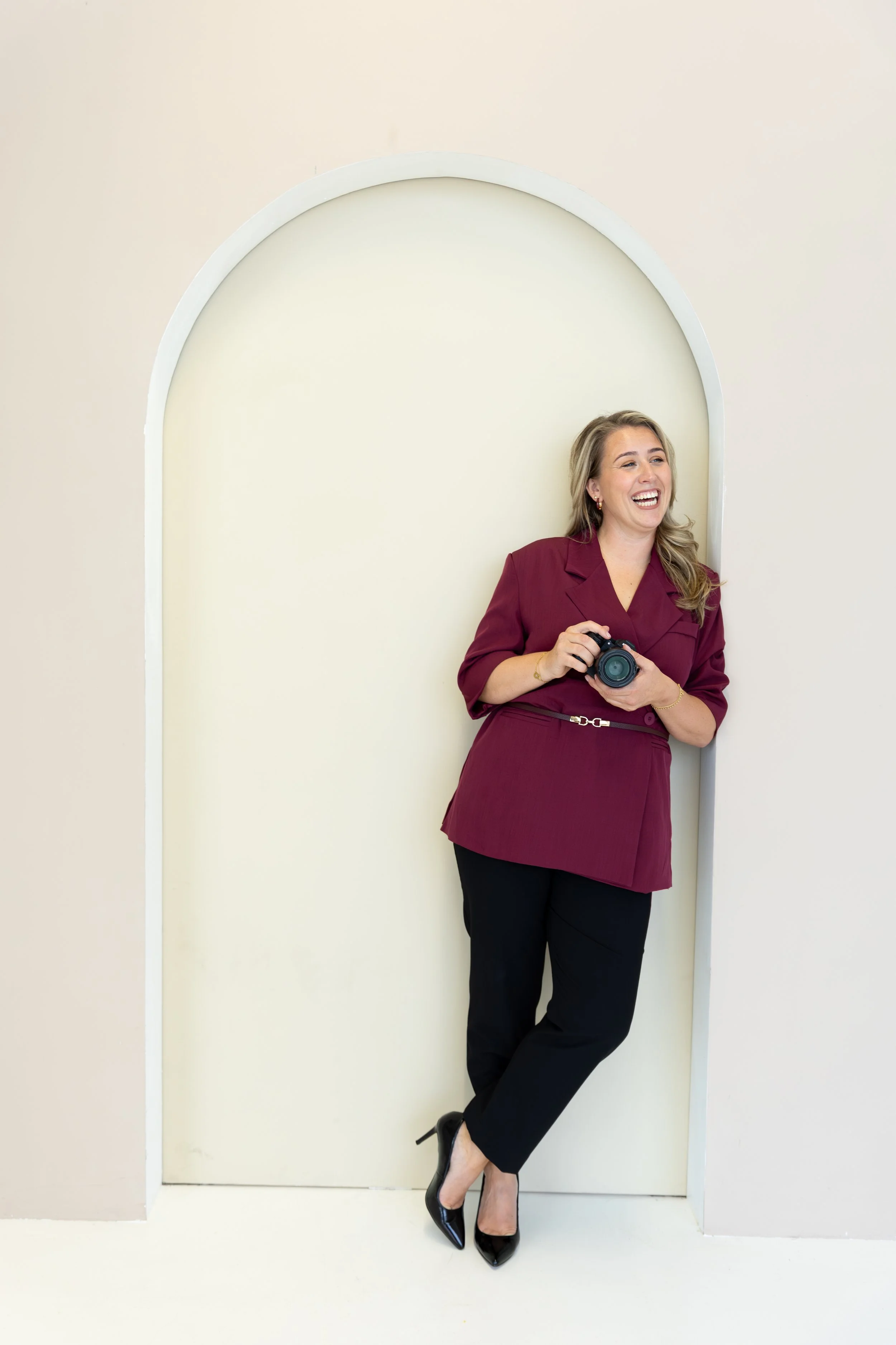 Vrouw in een bordeauxrode blazer en zwarte pantalons met hoge hiel schoenen, lachend met een camera in haar hand.