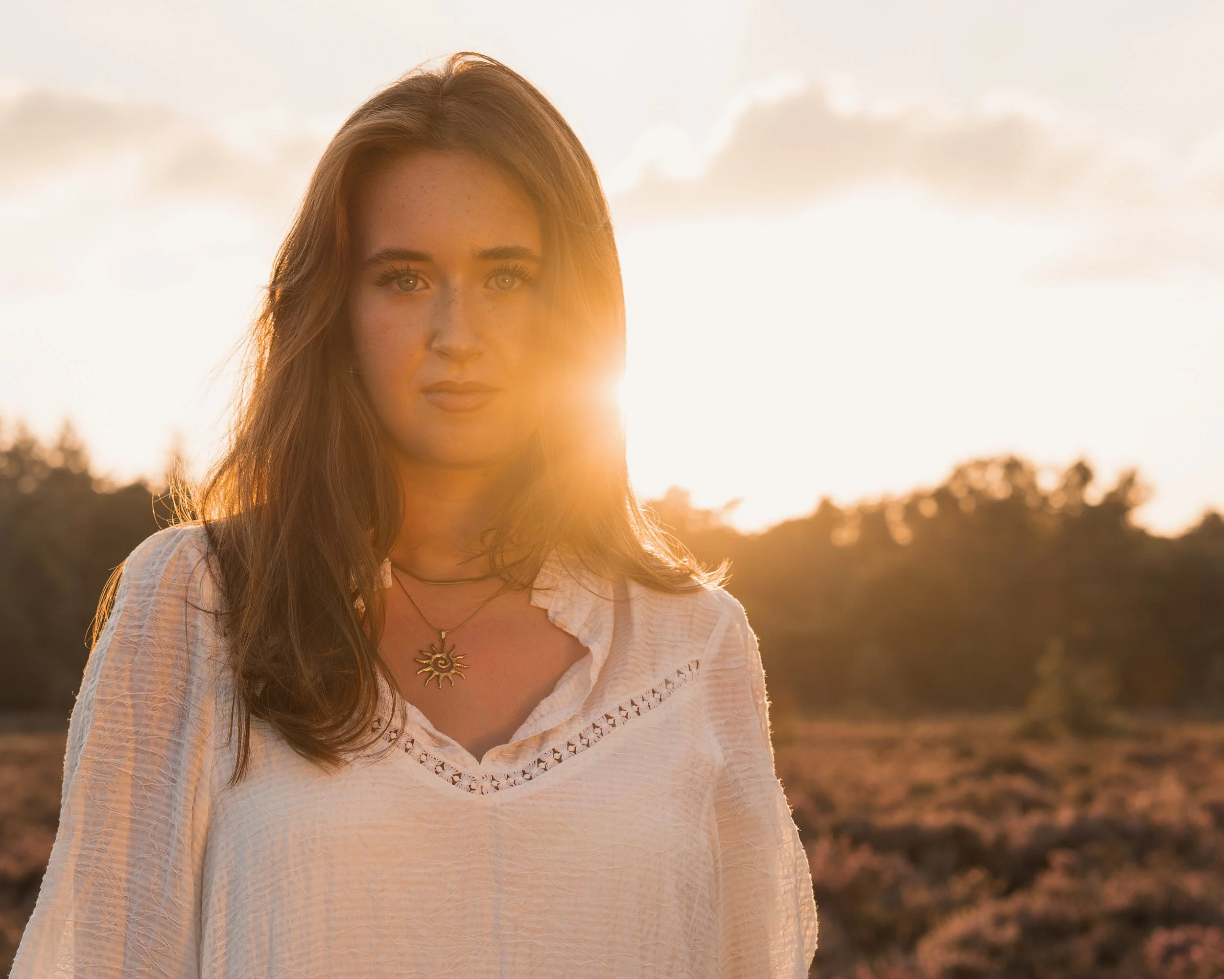 Portret van een jonge vrouw met bruin haar, witte blouse, en een zonne-edelsteen hanger, staand in een veld tijdens de zonsondergang.