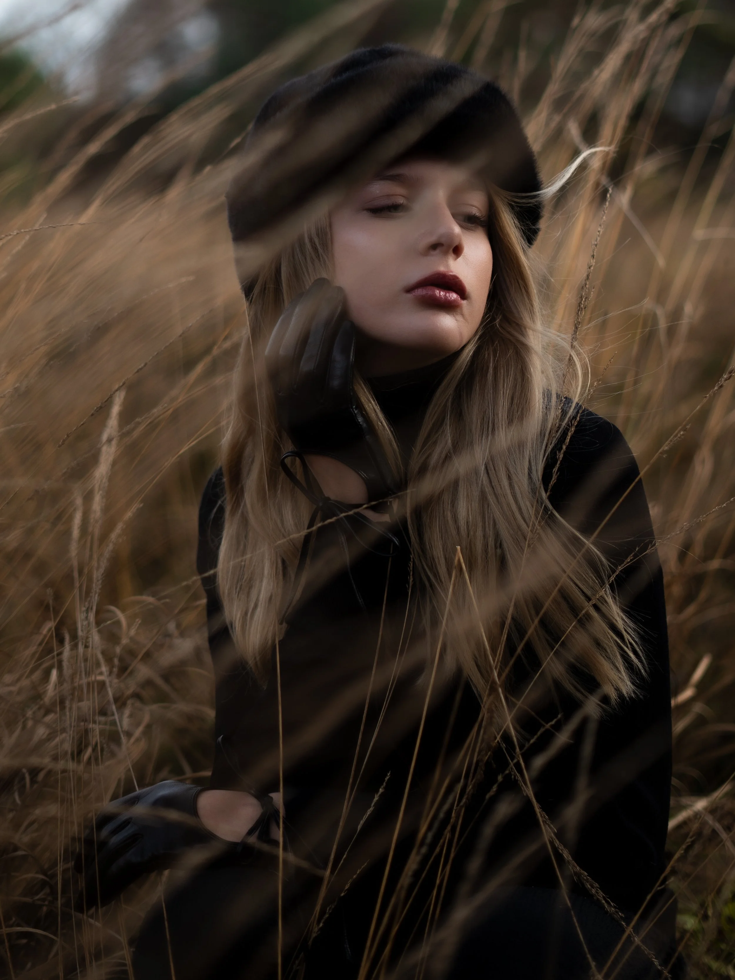 Een vrouw met lange, golvende haar draagt een zwarte hoed en handschoenen, zittend tussen droge grassen.