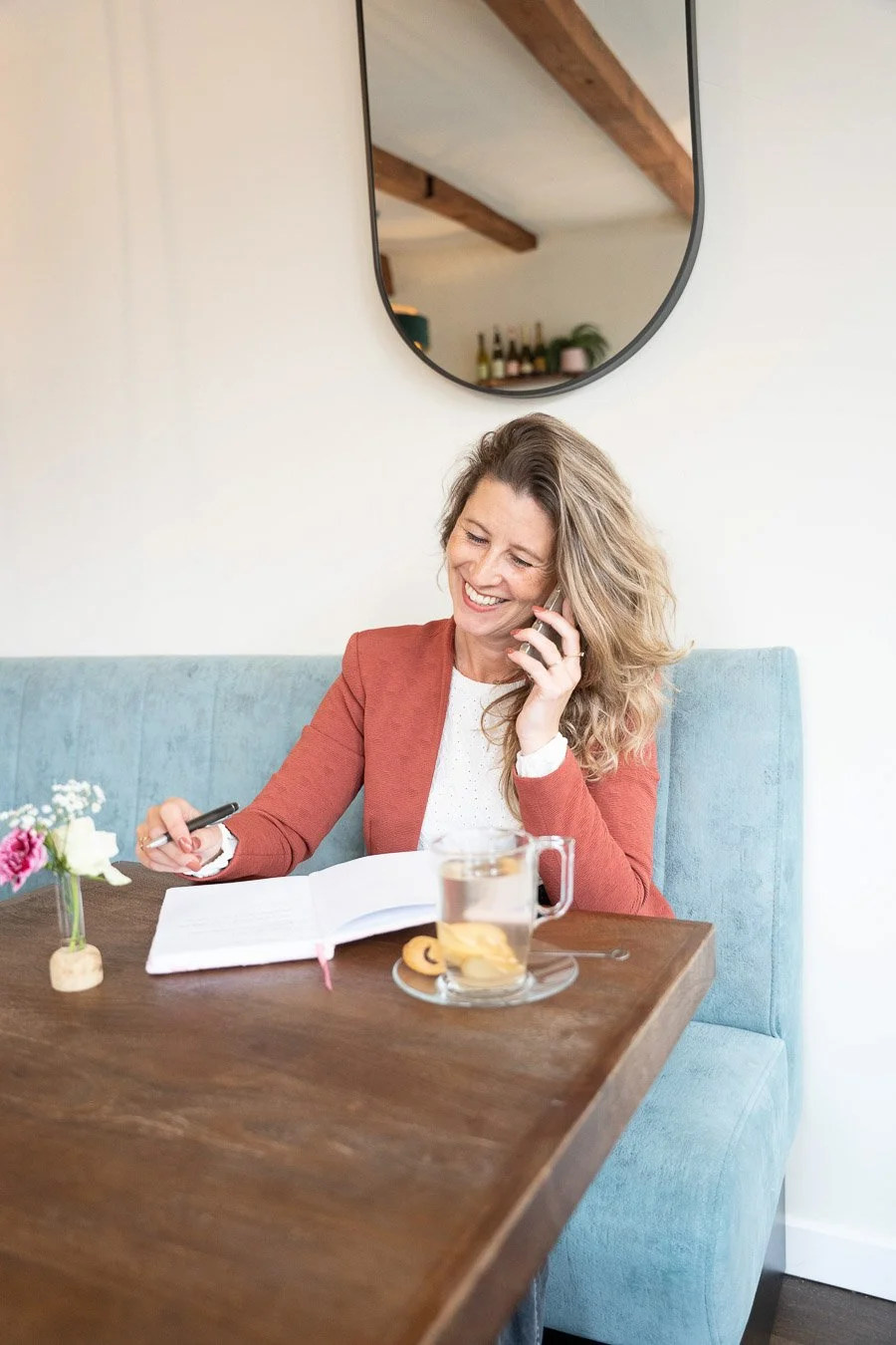 Vrouw zit aan een tafel met een open notitieboek, een pen, een glas thee met citroen en een koekje, ze belt en glimlacht.
