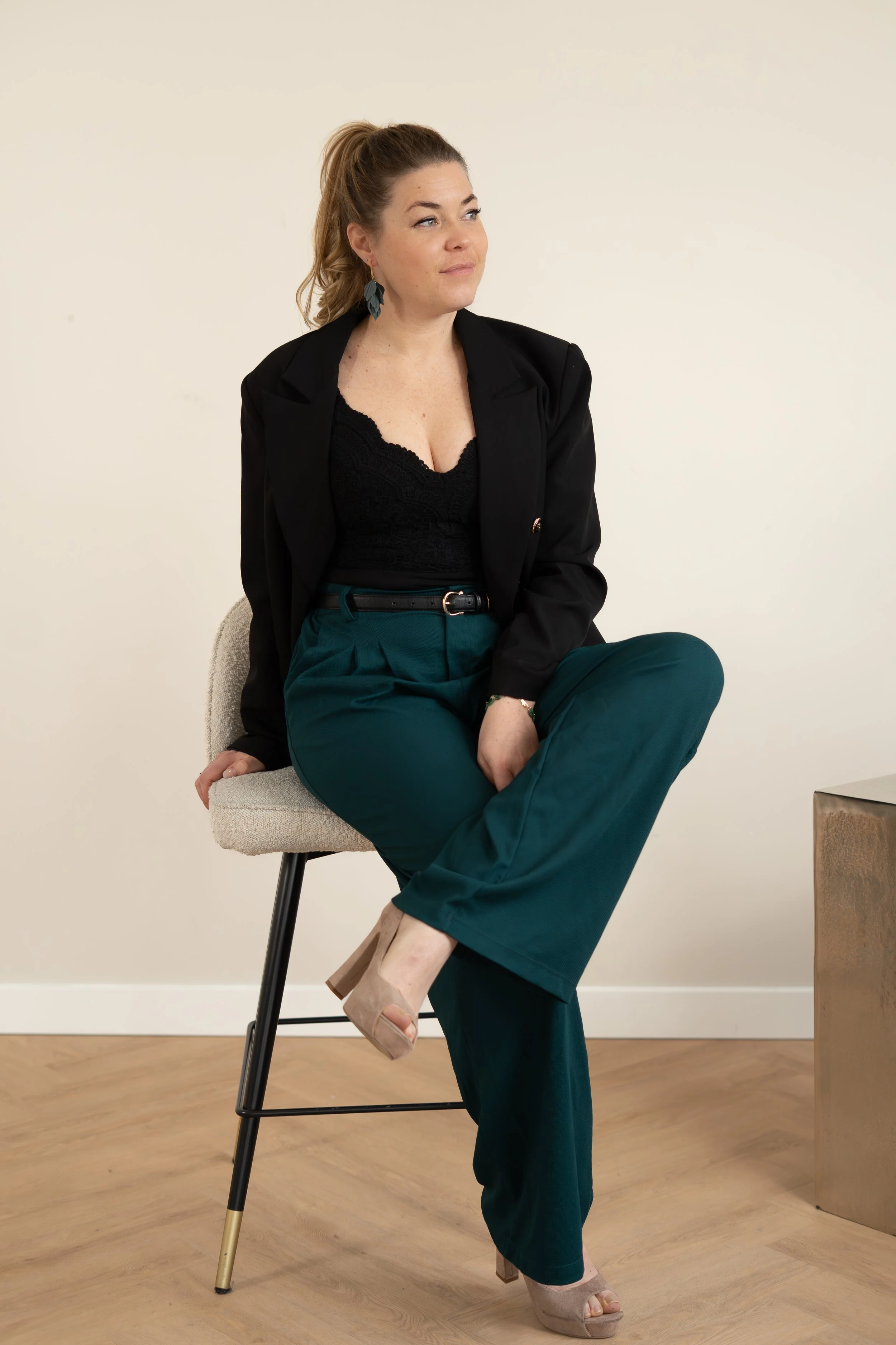 Vrouw zit op een kruk, gekleed in een zwarte blazer, zwarte top en groene broek, met een houding waarin ze haar benen gekruist heeft, in een kamergezellige omgeving.