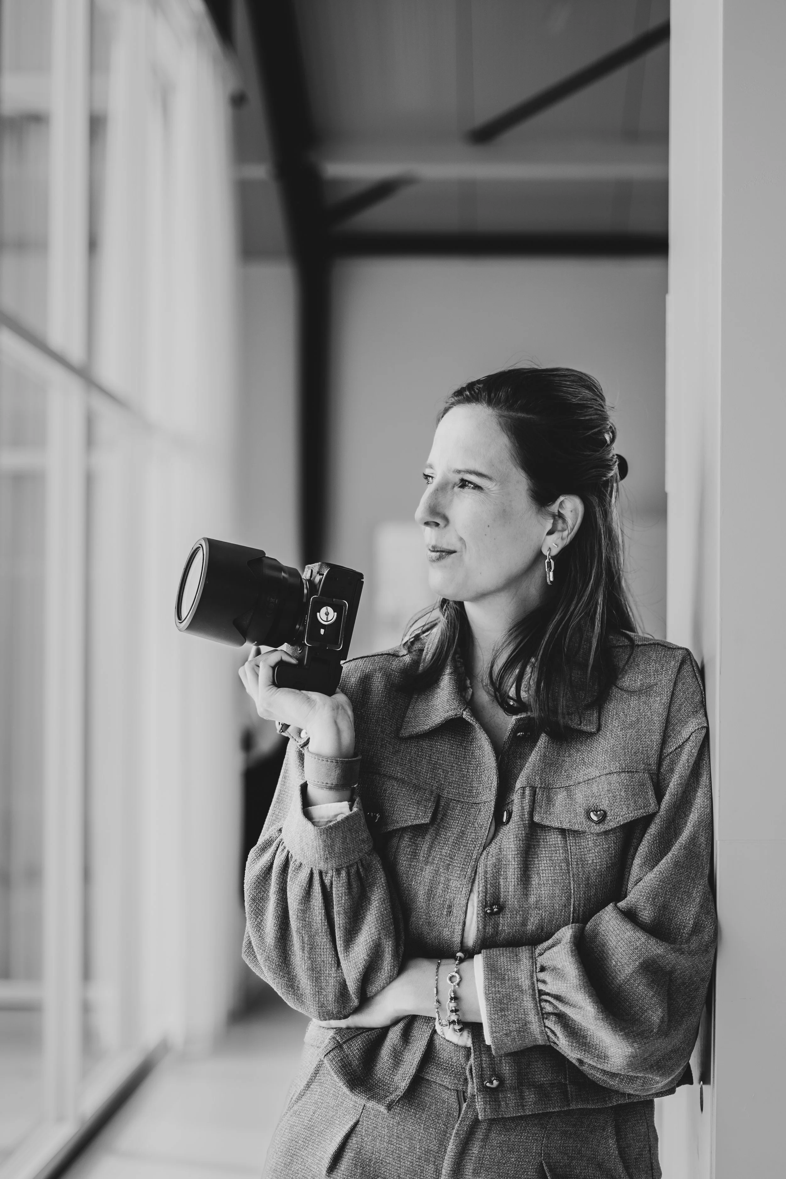 Een vrouw staat naast een raam en houdt een camera in haar rechterhand. Ze kijkt nadenkend naar buiten, draagt een colbert en sieraden.