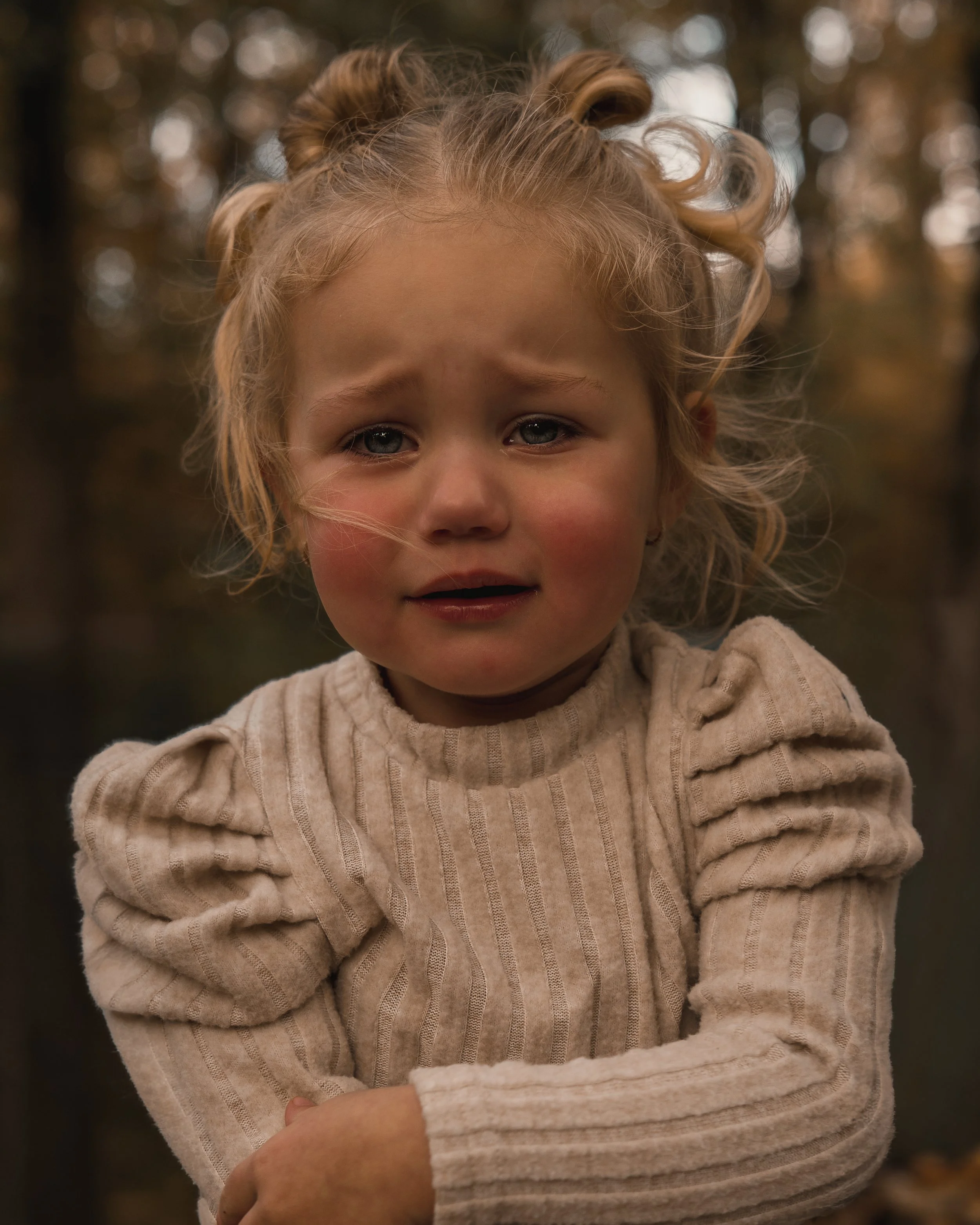 Een jonge meisje met blonde, gekrulde haar in een bos, met een uitdrukking van verdriet of verwarring op haar gezicht