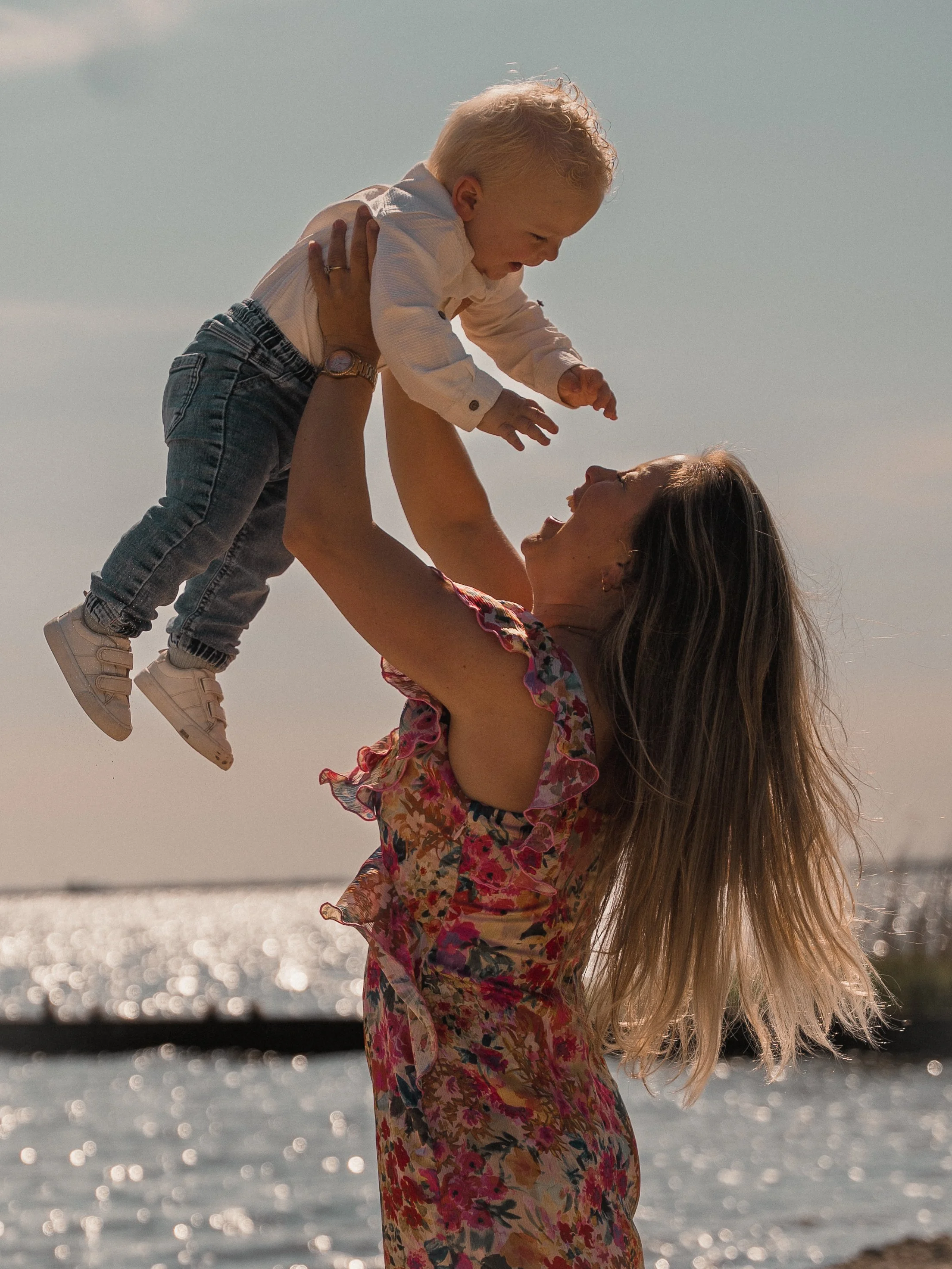 Een vrouw die haar kind op een pier bij het water omhoog houdt, lachend tot het kind, met de zonsondergang op de achtergrond.