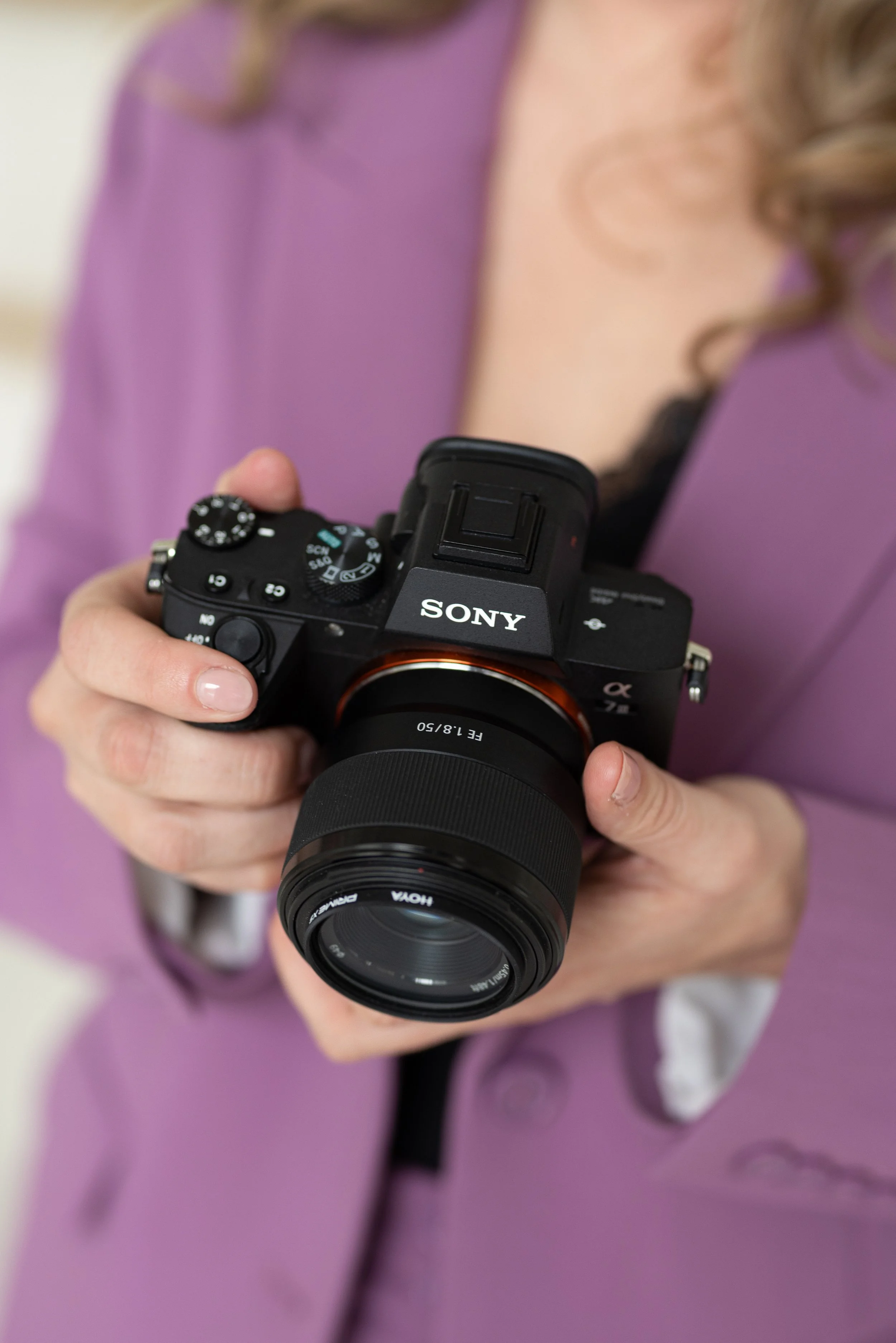 Dichtbij opname van een persoon die een Sony camera vasthoudt, de persoon draagt een paarse jas en heeft lichtgekleurde nagels.