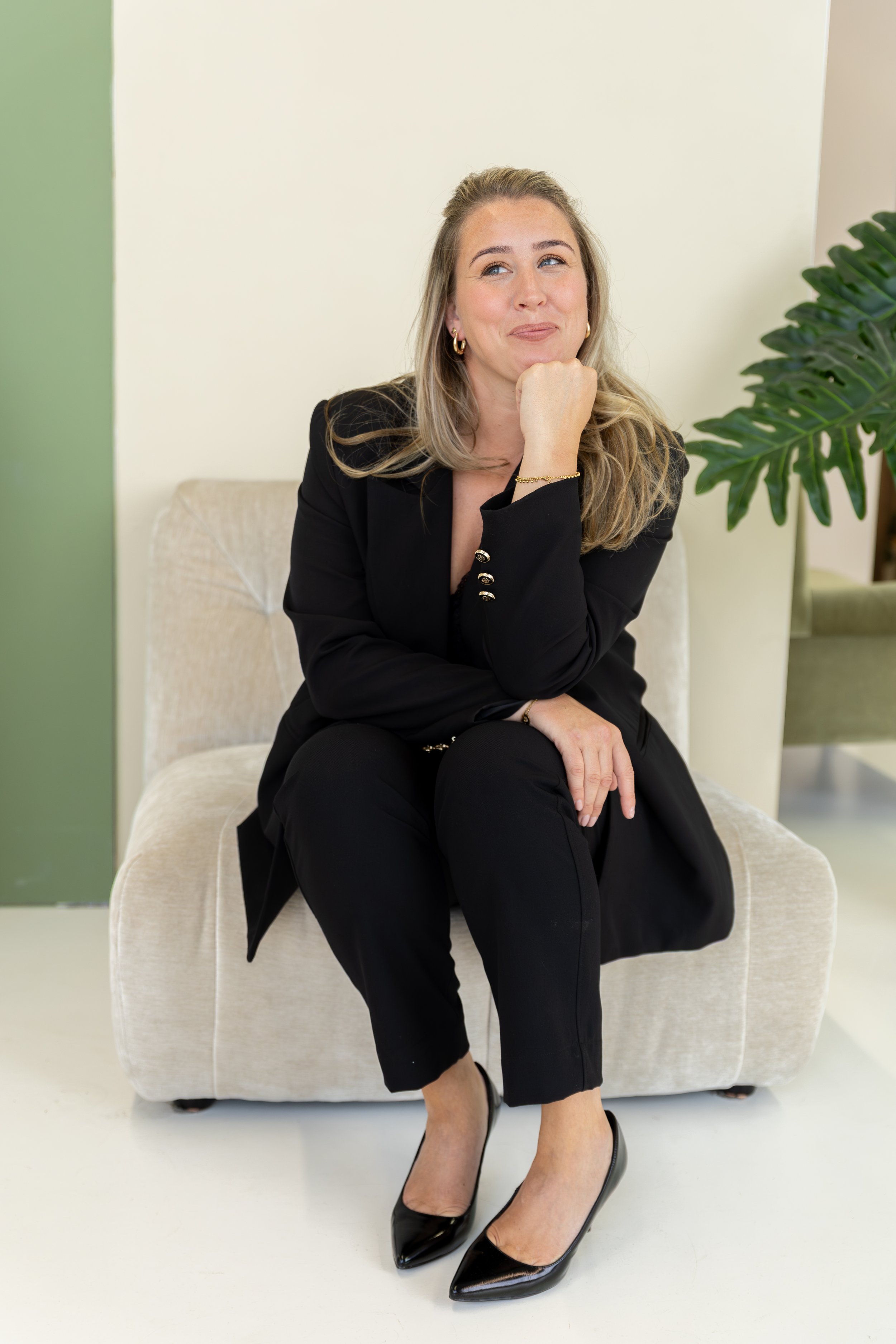 Vrouw in zwart pak zittend op een beige bank met een groene deur en een grote plant in de achtergrond.