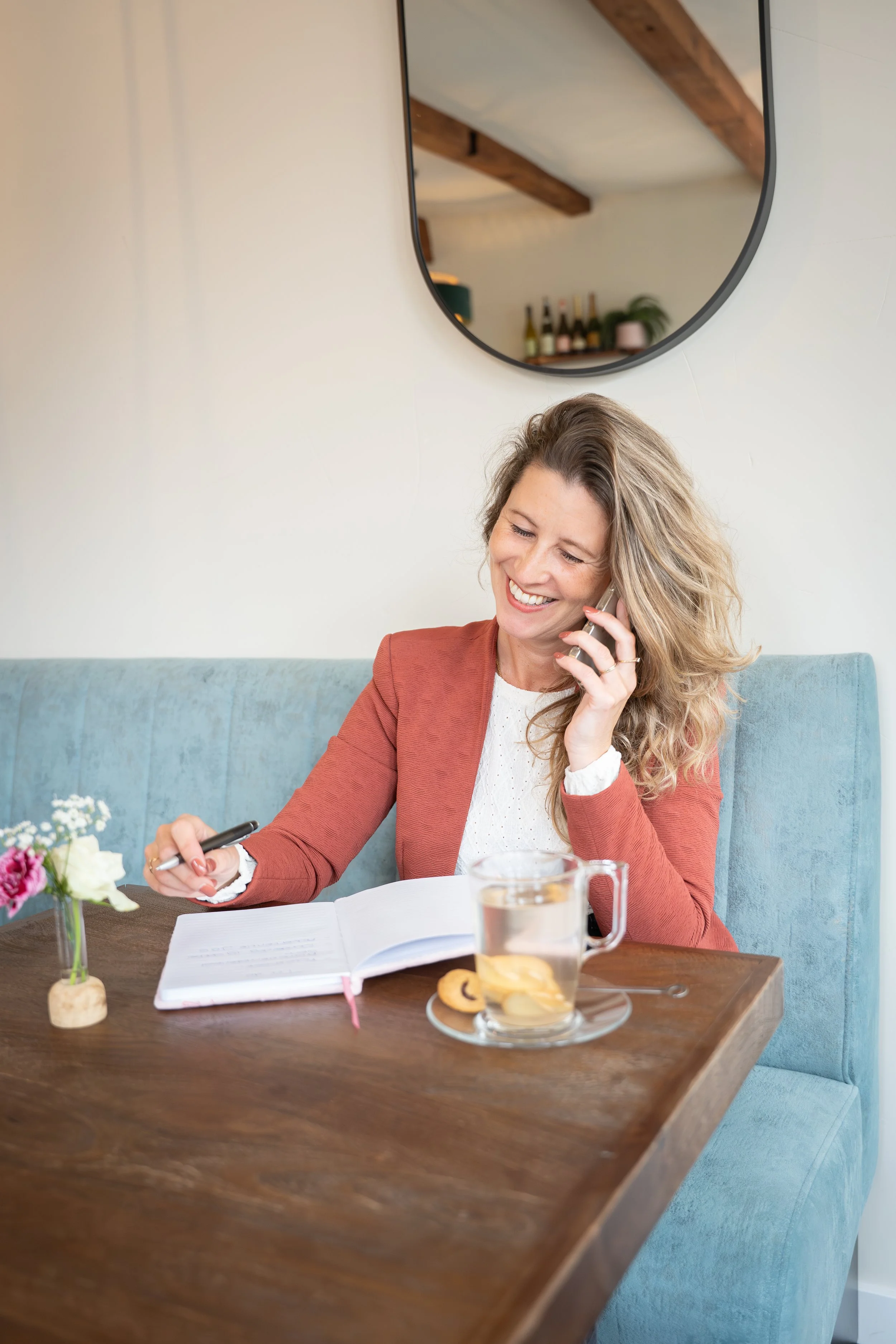 Vrouw zit aan een houten tafel, maakt aantekeningen in een open notitieboek, telefoneert en drinkt water met een koekje op een schotel.
