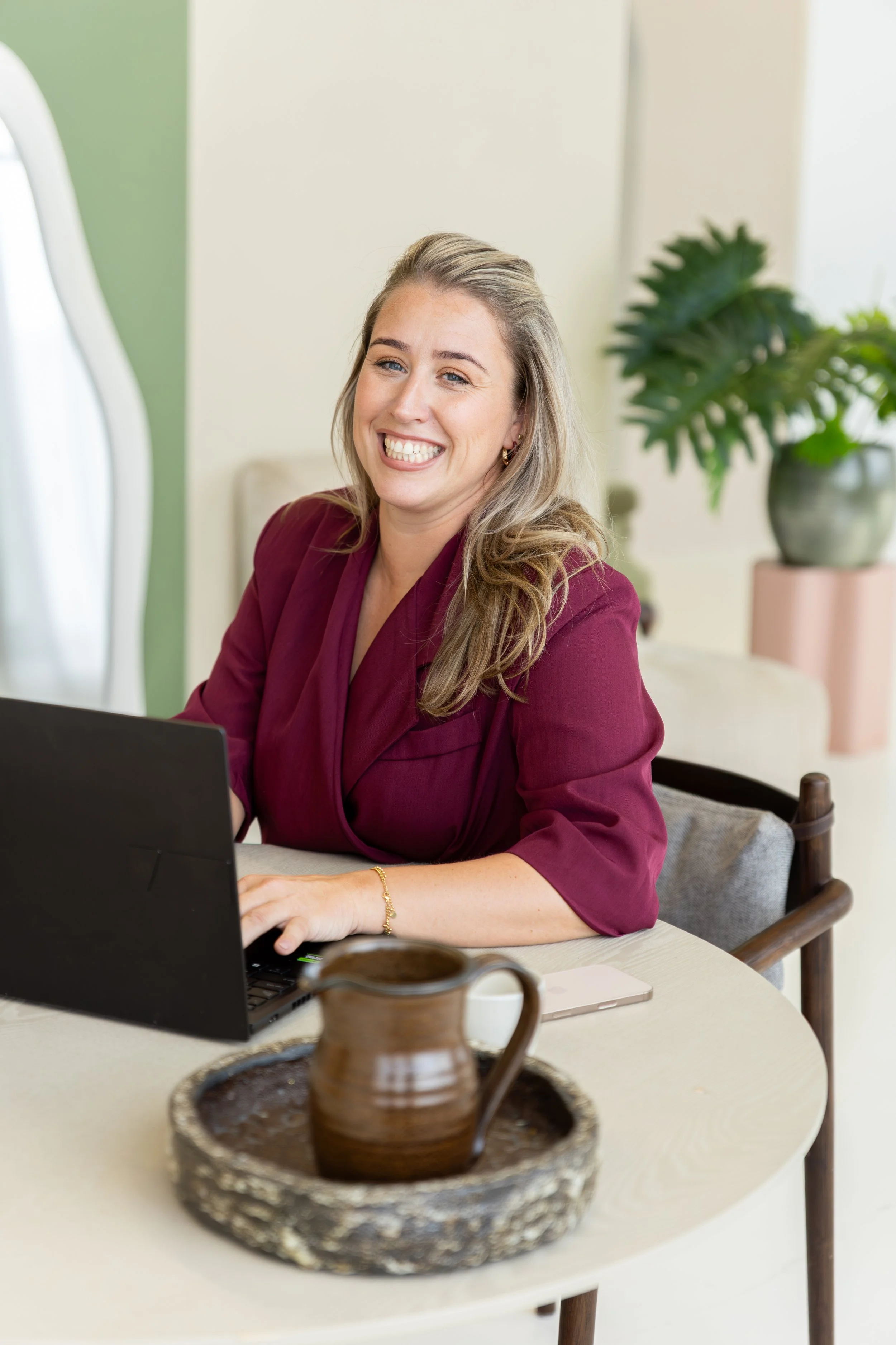 Een glimlachende vrouw in een bordeauxrode blouse zit aan een tafel met een laptop, een kopje en een smartphone, met een groene plant op de achtergrond.