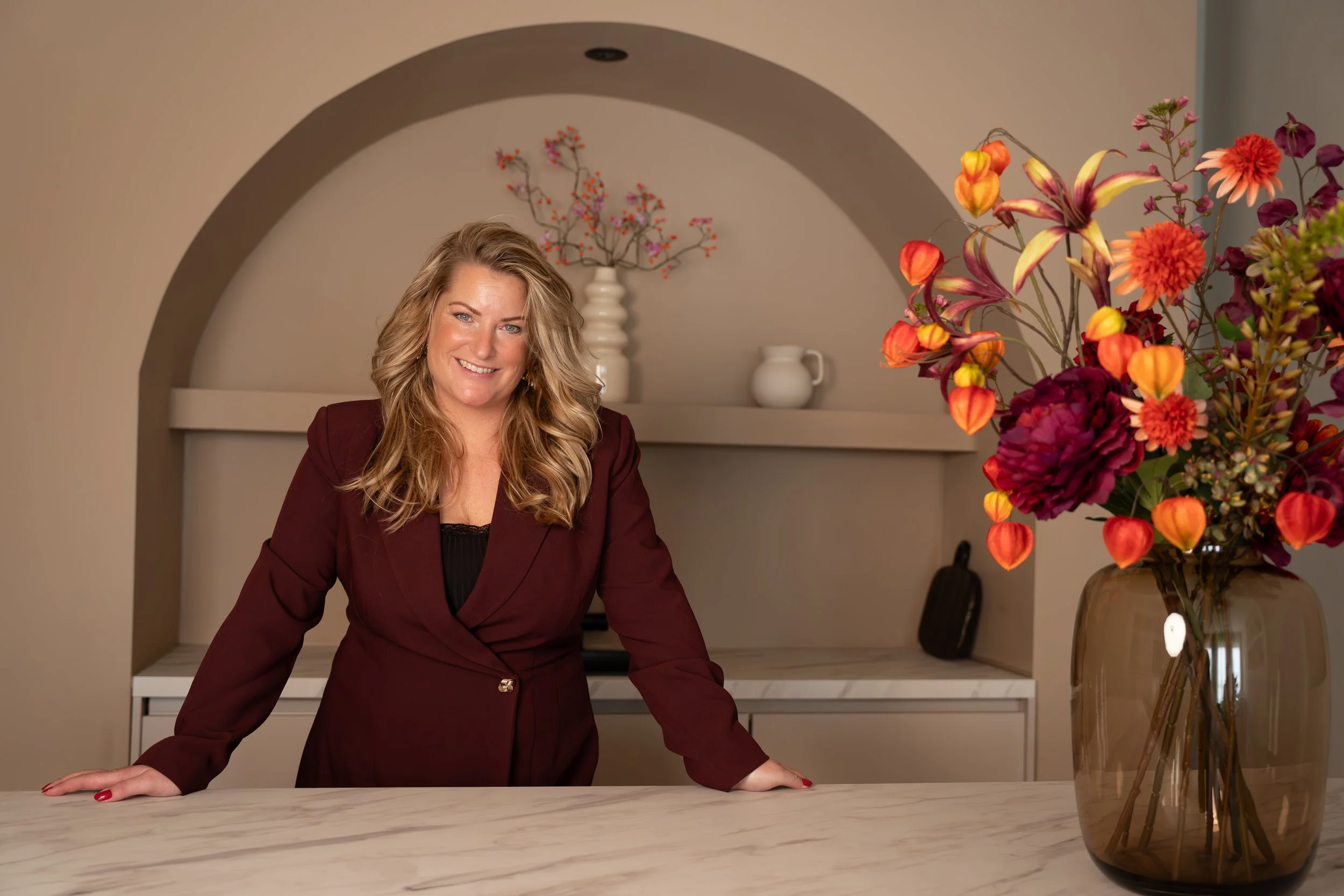 Een glimlachende vrouw met lang, golfachtig haar, gekleed in een donkere roodbruine jas, leunt op een marmeren keukenblad met een grote vaas met kleurrijke bloemen en een achtergrond met decoratieve witte vazen.