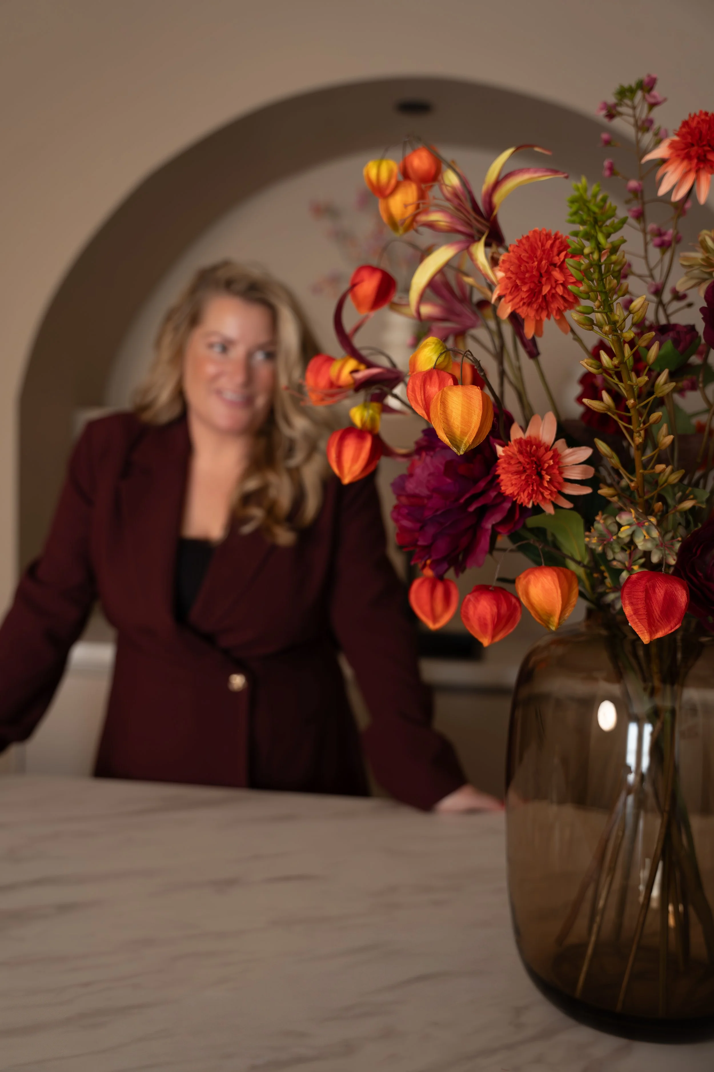 Close-up van een bloemenarrangement met roze en oranje bloemen in een glazen vaas, met een vrouw op de achtergrond die naar de bloemen kijkt.