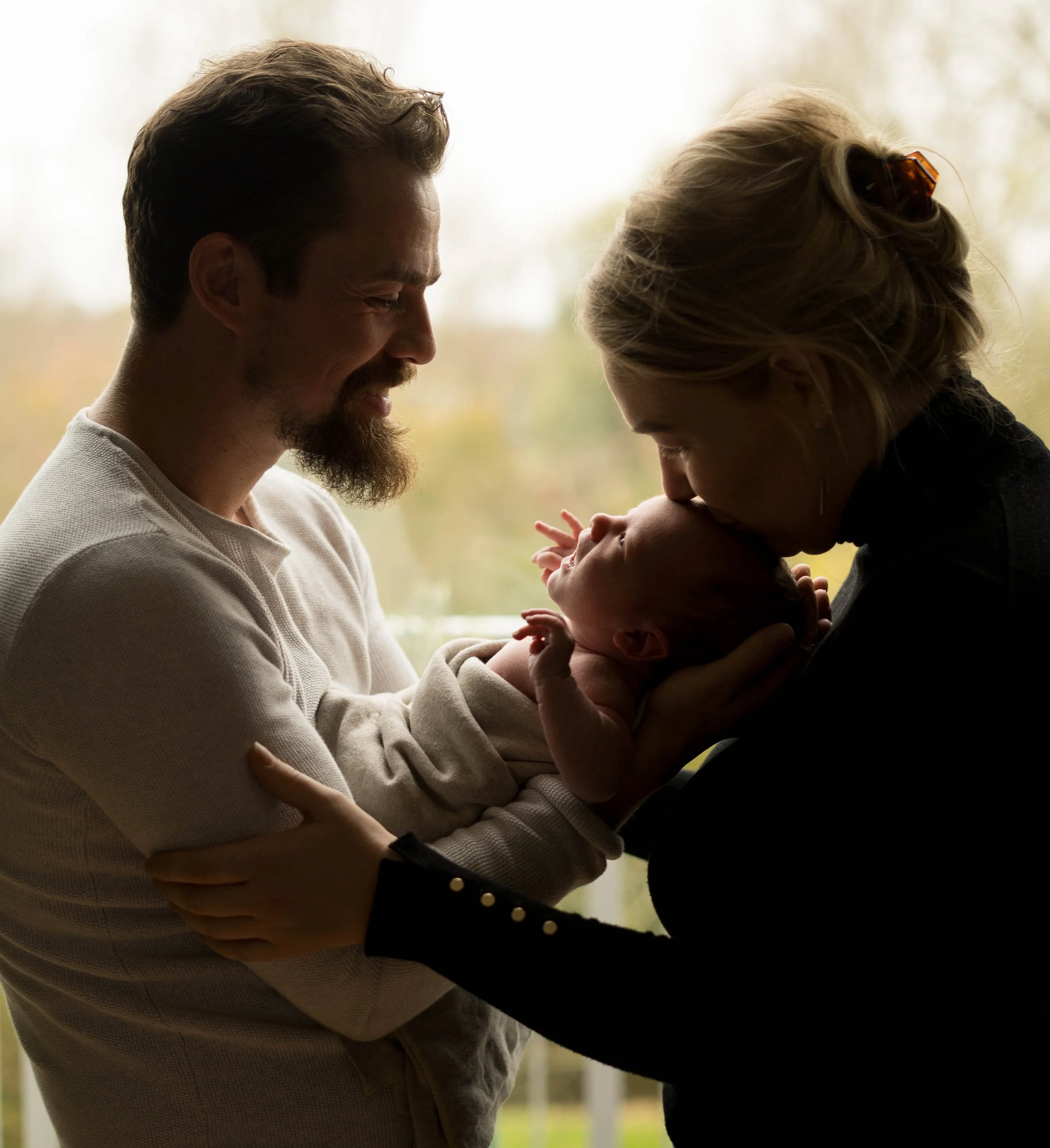 Een man en een vrouw houden een pasgeboren baby vast; de vrouw kust de baby op het voorhoofd, met een zachte en liefdevolle uitdrukking. De achtergrond is vaag met natuurlijke buitenomgeving.