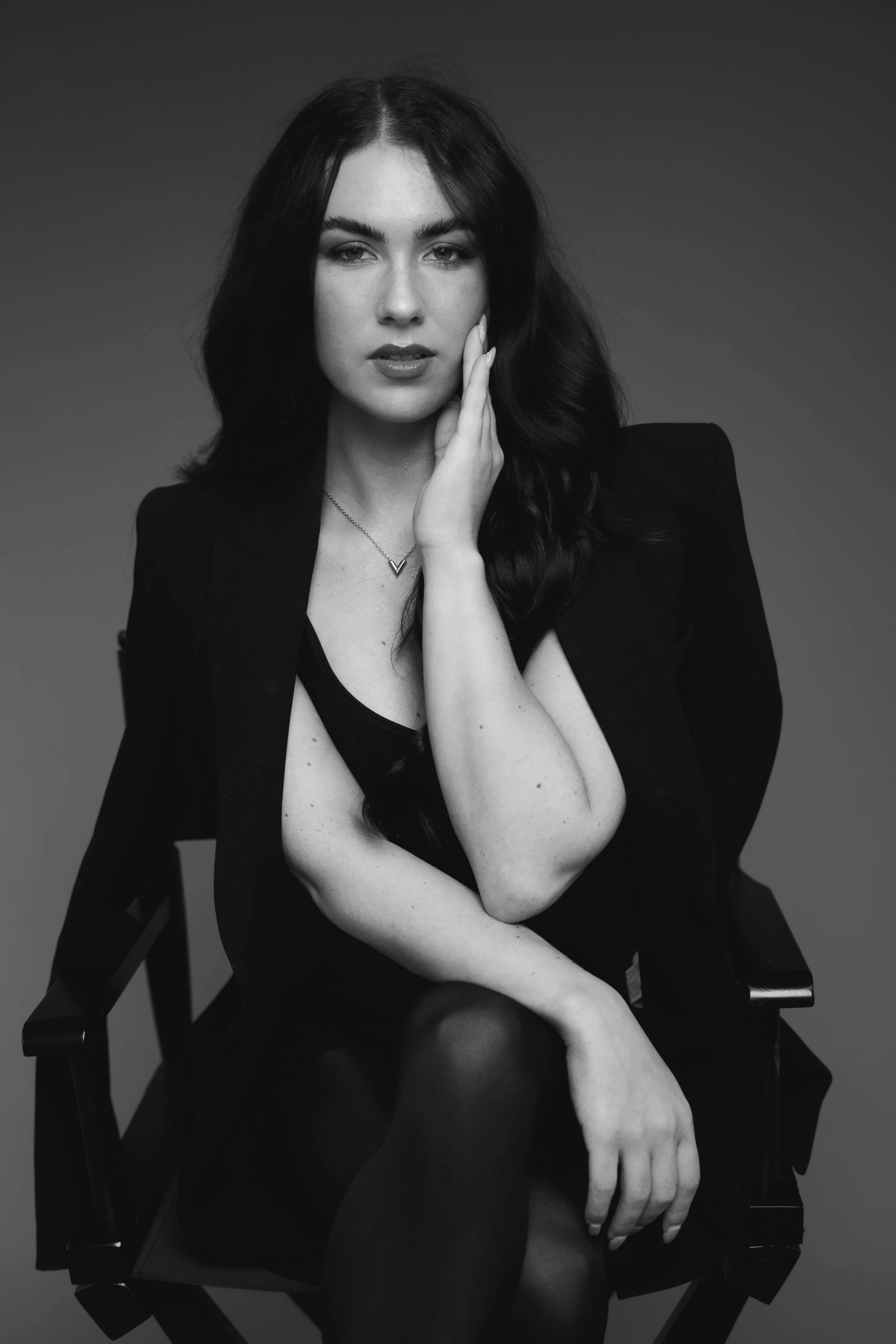 Zwarte en witte portretfoto van een vrouw met lang, golvend haar die zit op een stoel, haar hand op haar gezicht en een serieuze uitdrukking, donkere achtergrond.