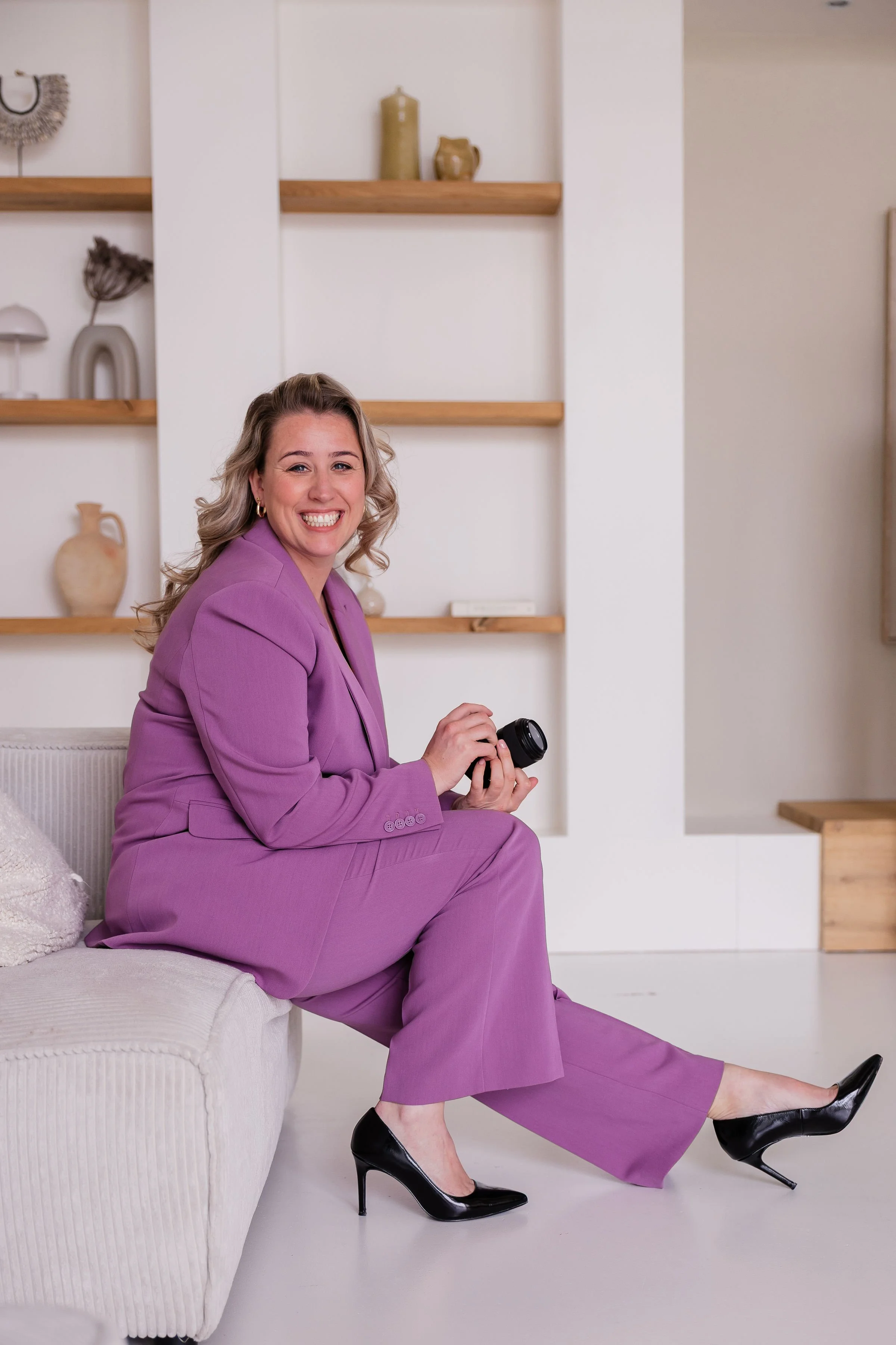 Vrouw in een paarse pak met hoge hakken zit op een bankje, lacht en houdt een camera vast.