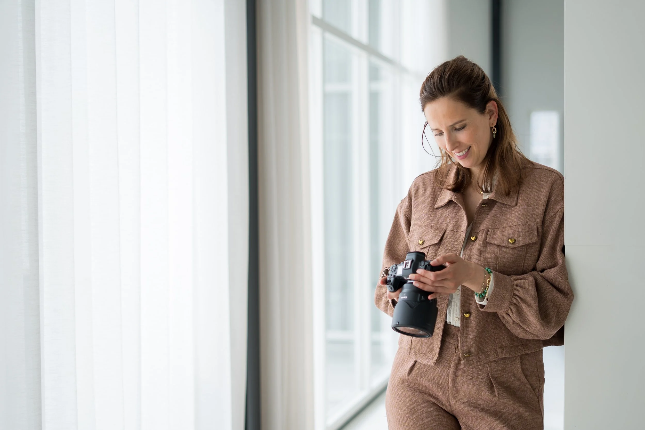Vrouw die een camera vasthoudt en naar de camera glimlacht in een lichte kamer met grote ramen.
