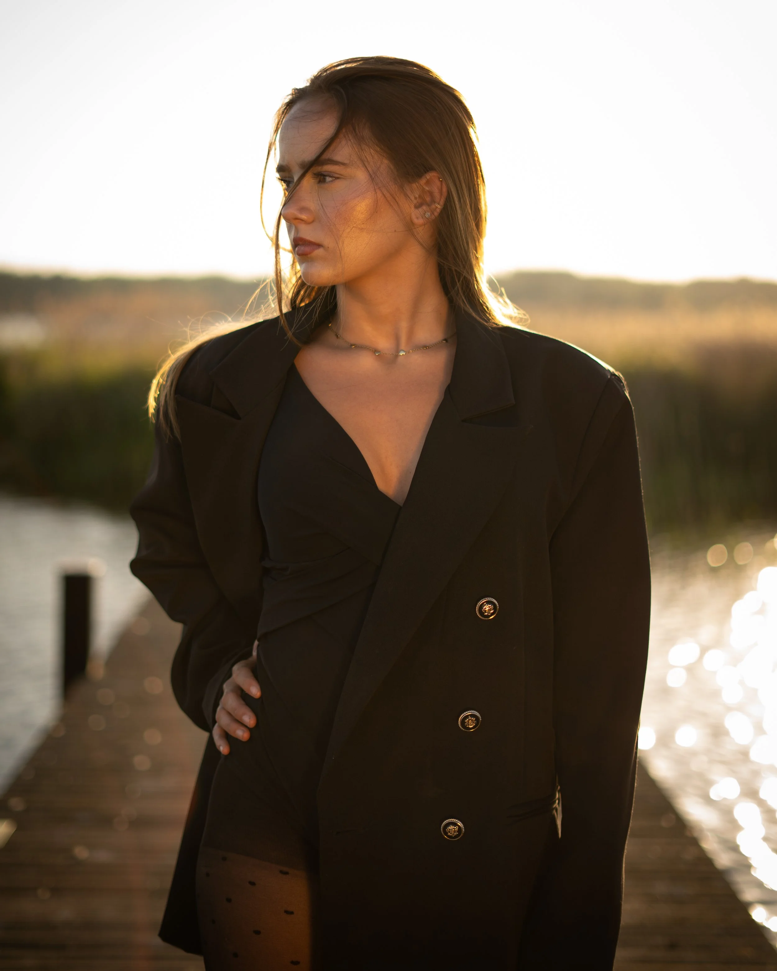 Een vrouw in een zwarte jas staat op een houten pier bij het water tijdens zonsondergang.